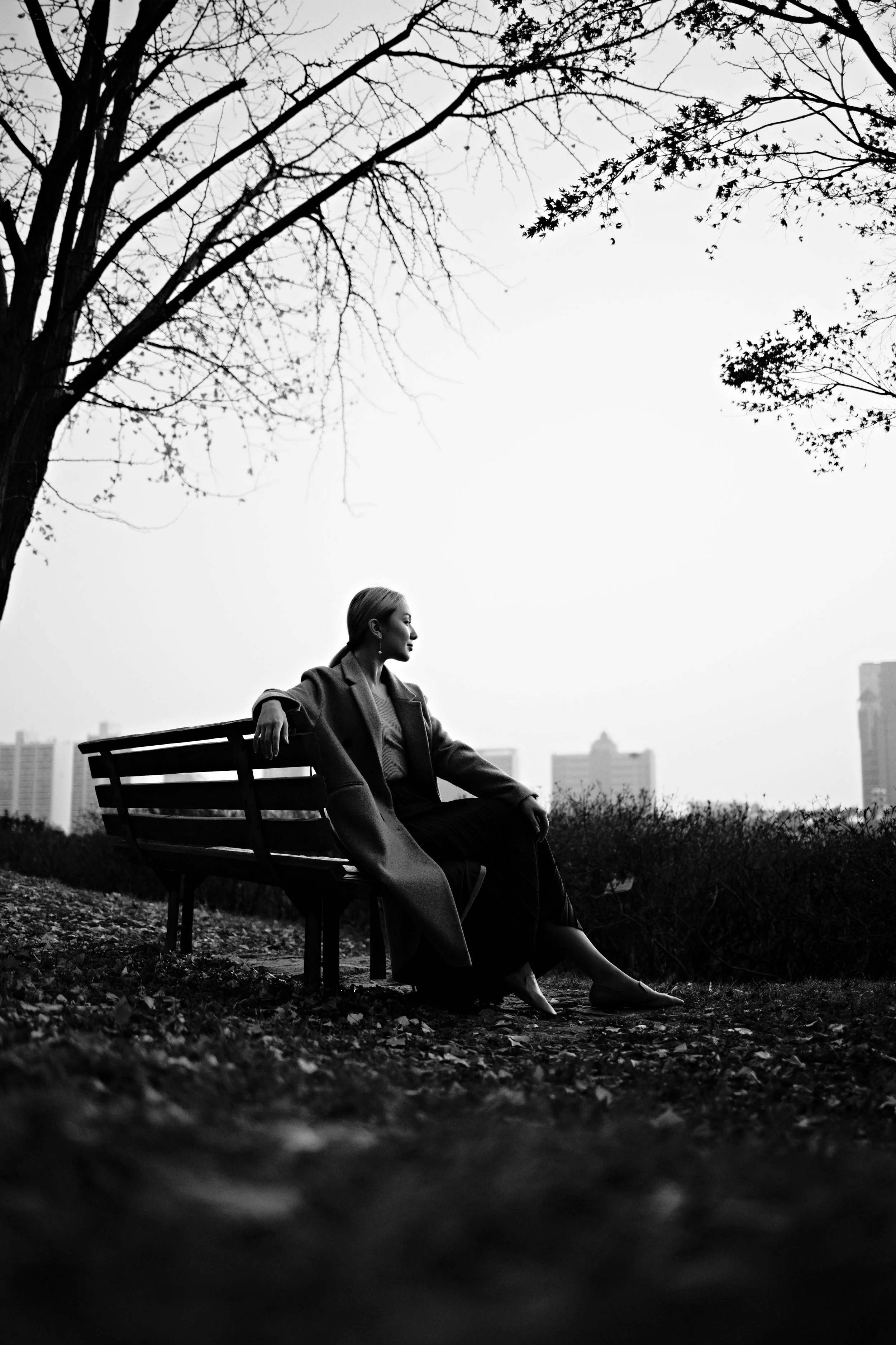 A woman sitting on a park bench, looking to the side, with trees and city buildings in the background, captured in black and white.