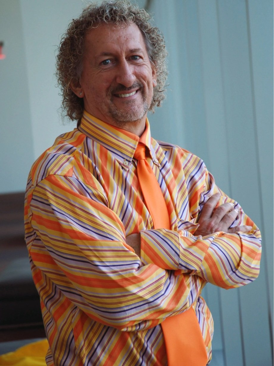 A man with curly gray hair and a beard wearing a colorful, striped shirt with orange, yellow, and purple tones, and an orange tie, standing with arms crossed and smiling in front of a blue wooden wall.