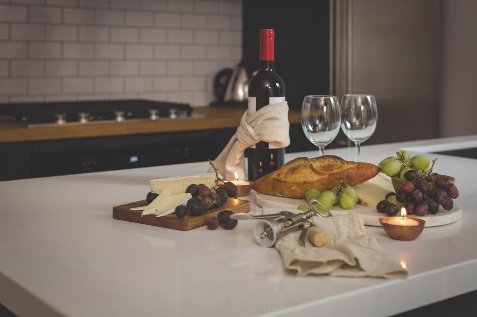 A kitchen counter with a bottle of red wine wrapped in a cloth, two empty wine glasses, cheese, grapes, a loaf of bread, a candle, and wine opener on a cloth.