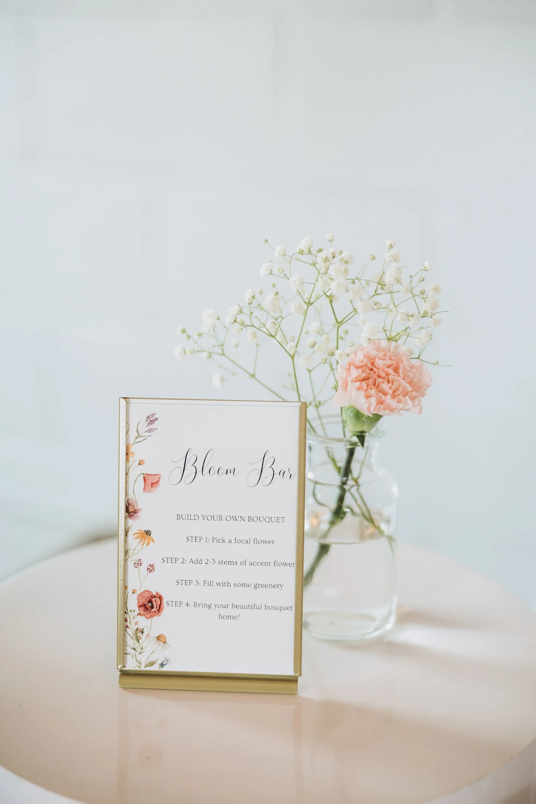 A sign for a 'Bloom Bar' with instructions to build a bouquet, placed on a round table next to a glass jar with pink carnations and white baby's breath flowers.