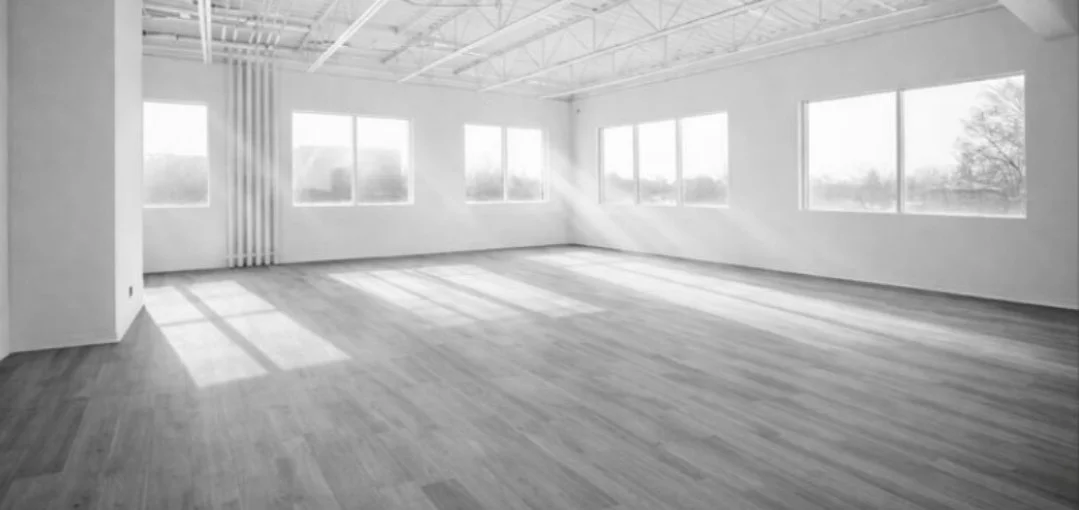 Empty room with large windows, hardwood floors, and white walls, sunlight casting shadows on the floor.