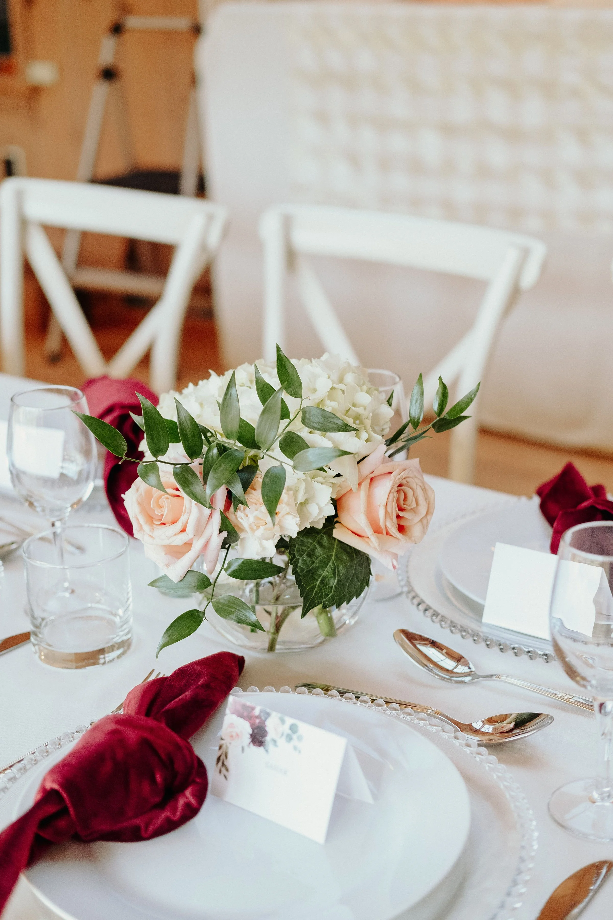 Elegant table setting with a floral centerpiece, including pale pink roses, white hydrangeas, and green leaves, surrounded by glassware, plates, and silverware for a formal event.