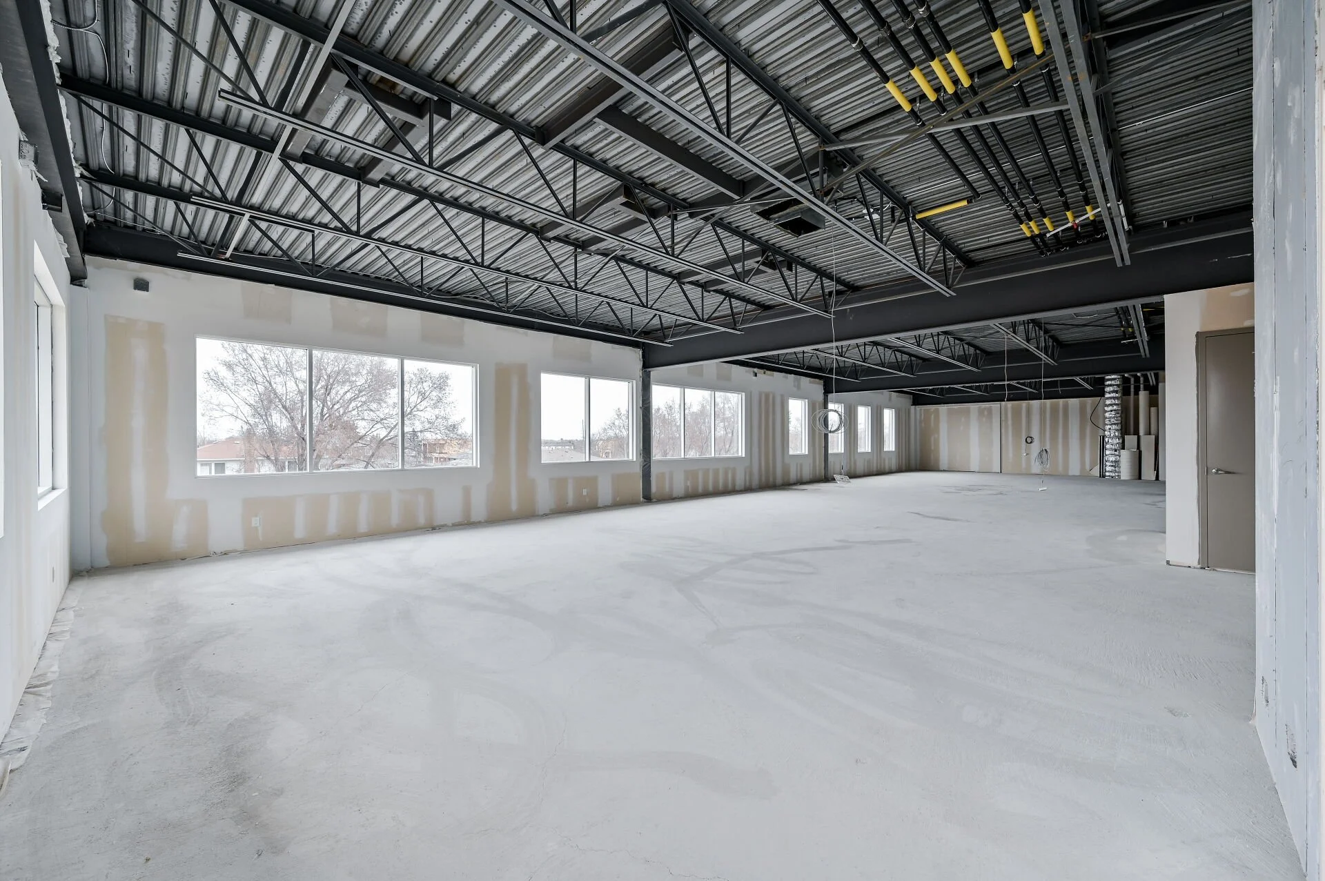 An unfinished interior space under construction with large windows, exposed ceiling with black metal framework, and white unfinished walls, with some construction materials and equipment in the background.