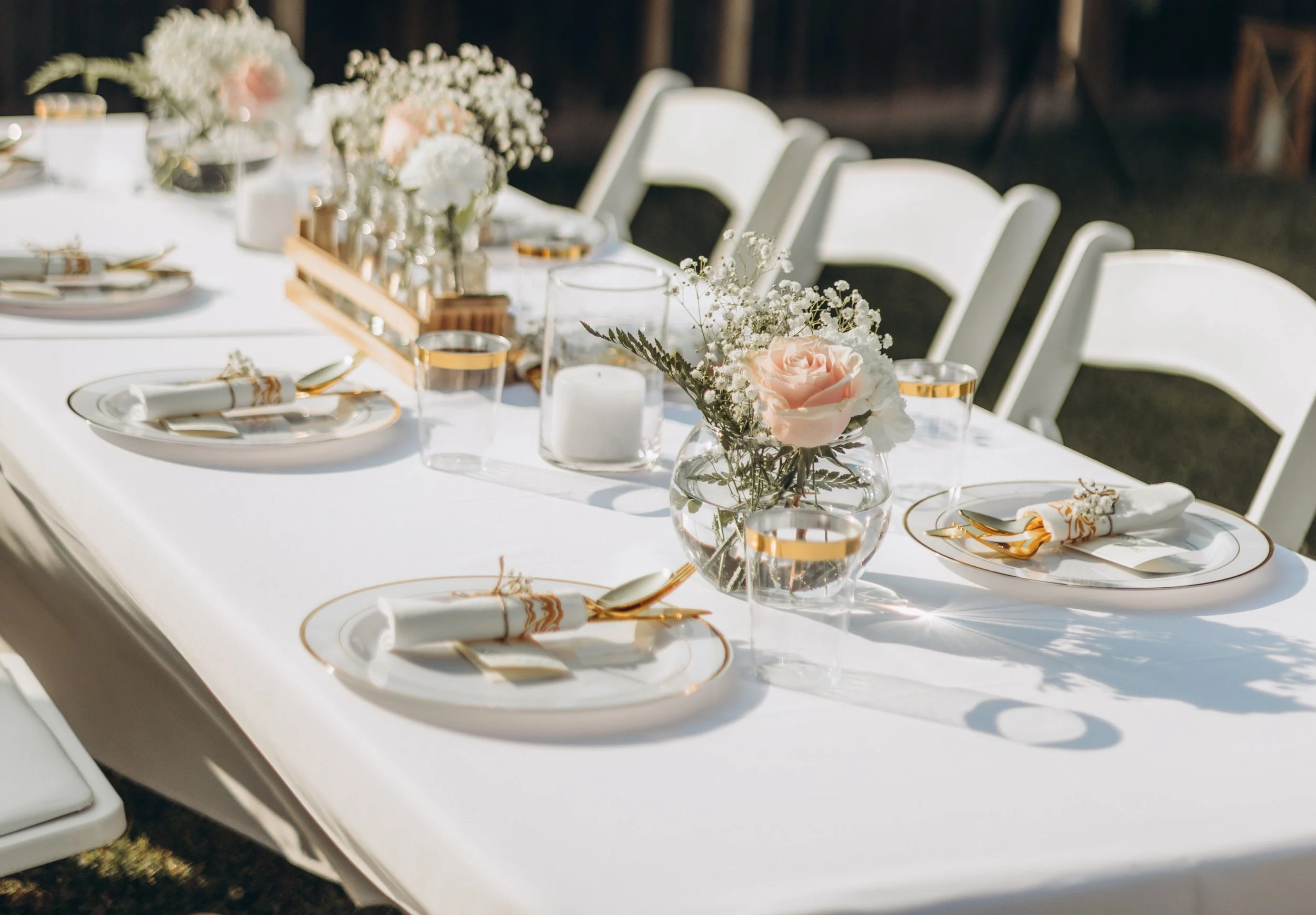 Elegant outdoor wedding table setup with white tablecloth, floral centerpieces with roses and baby's breath, candles, and gold-accented tableware.