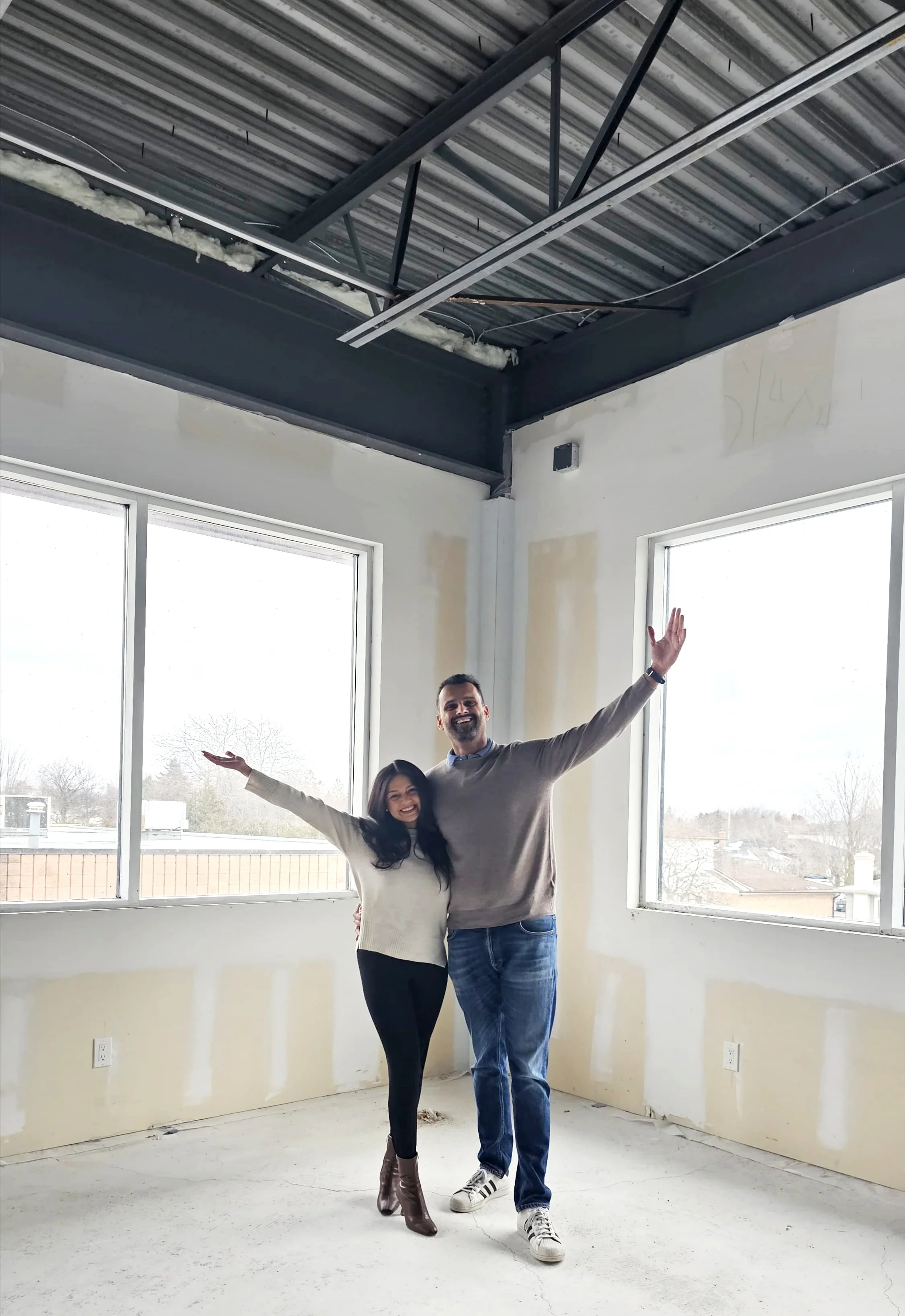 A smiling couple standing inside an unfinished room with exposed ceiling and drywall, with large windows showing a cloudy sky and neighborhood outside.