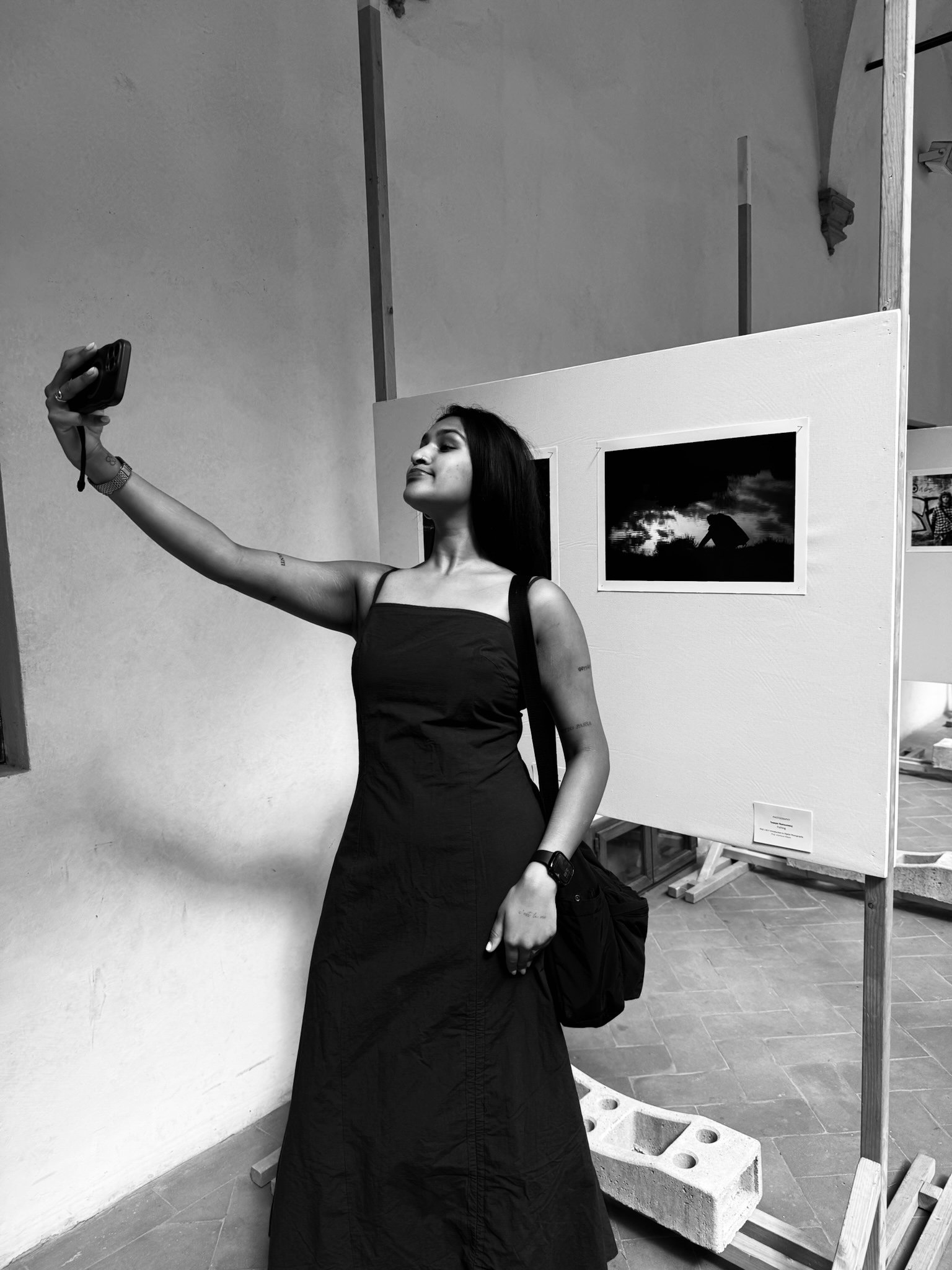 A woman in a black dress takes a selfie with her phone, standing next to an art display in a gallery or museum, with two framed photographs visible in the background.