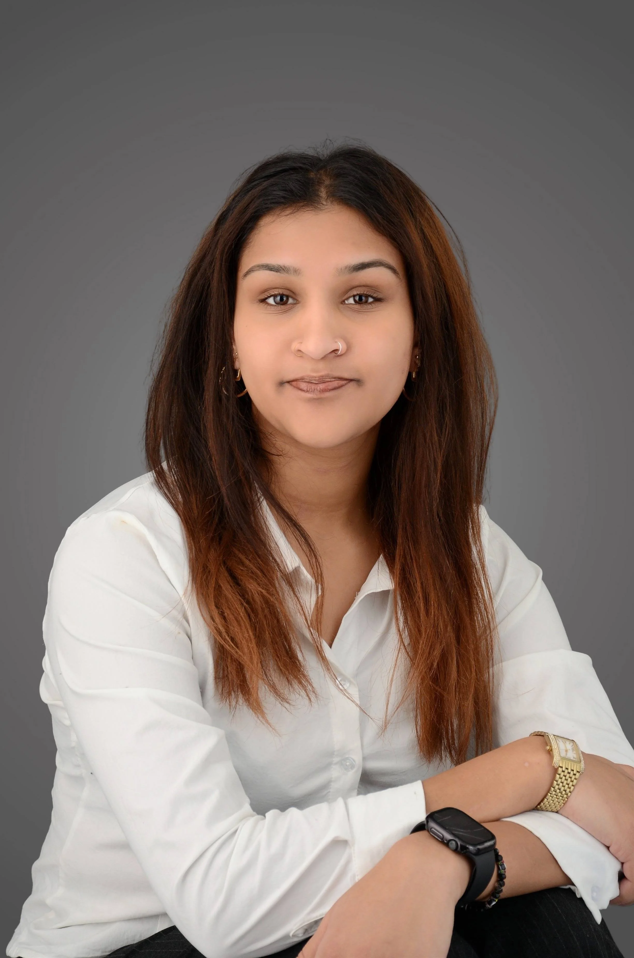 Portrait of a young woman with long, wavy brown hair, wearing a white shirt, gold watch, bracelets, and small earrings, sitting against a plain gray background.
