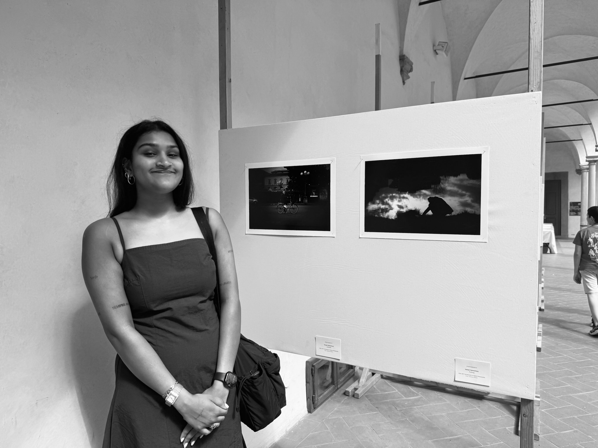 A young woman with dark hair, wearing earrings and a dark dress, standing indoors near a white display board with photographs. She has a slight smile and is standing with her arms crossed. The background shows arched ceilings and another person walking away.