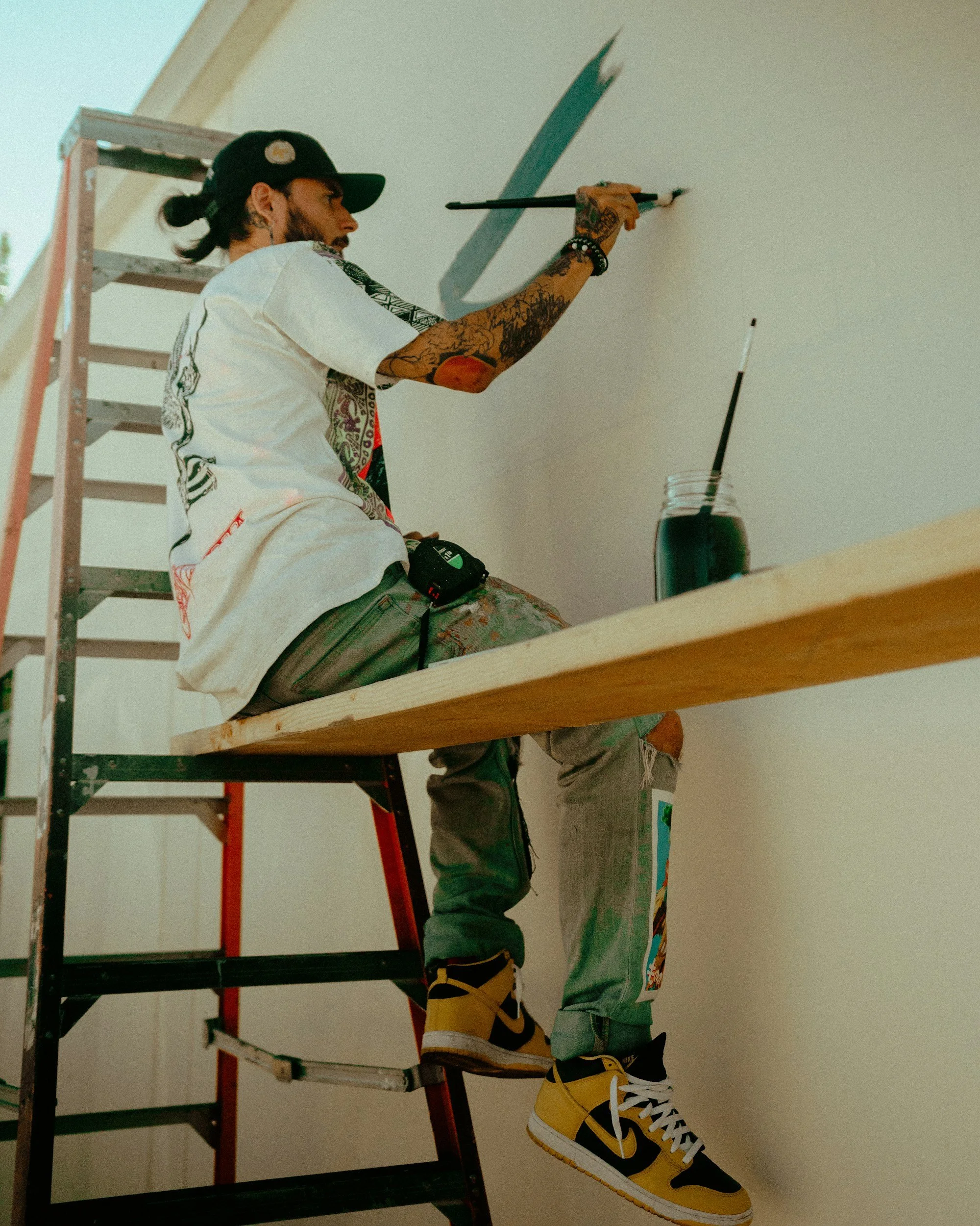 A man with tattoos wearing a black cap, white T-shirt, and yellow and black sneakers, sitting on a ladder and painting a wall with a brush.