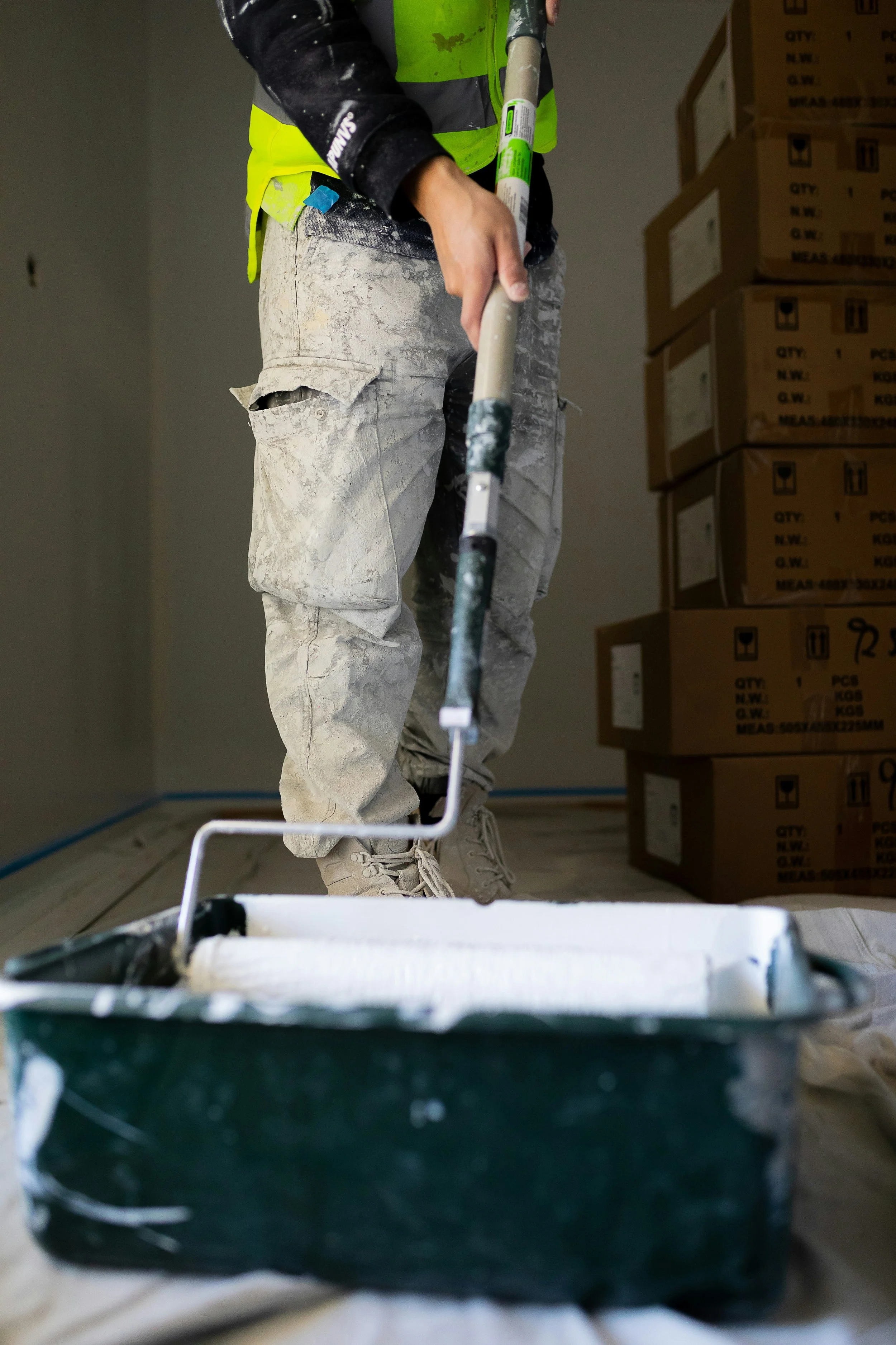 A person painting a wall with a paint roller, wearing work clothes and a safety vest, in a room with boxes stacked against the wall.