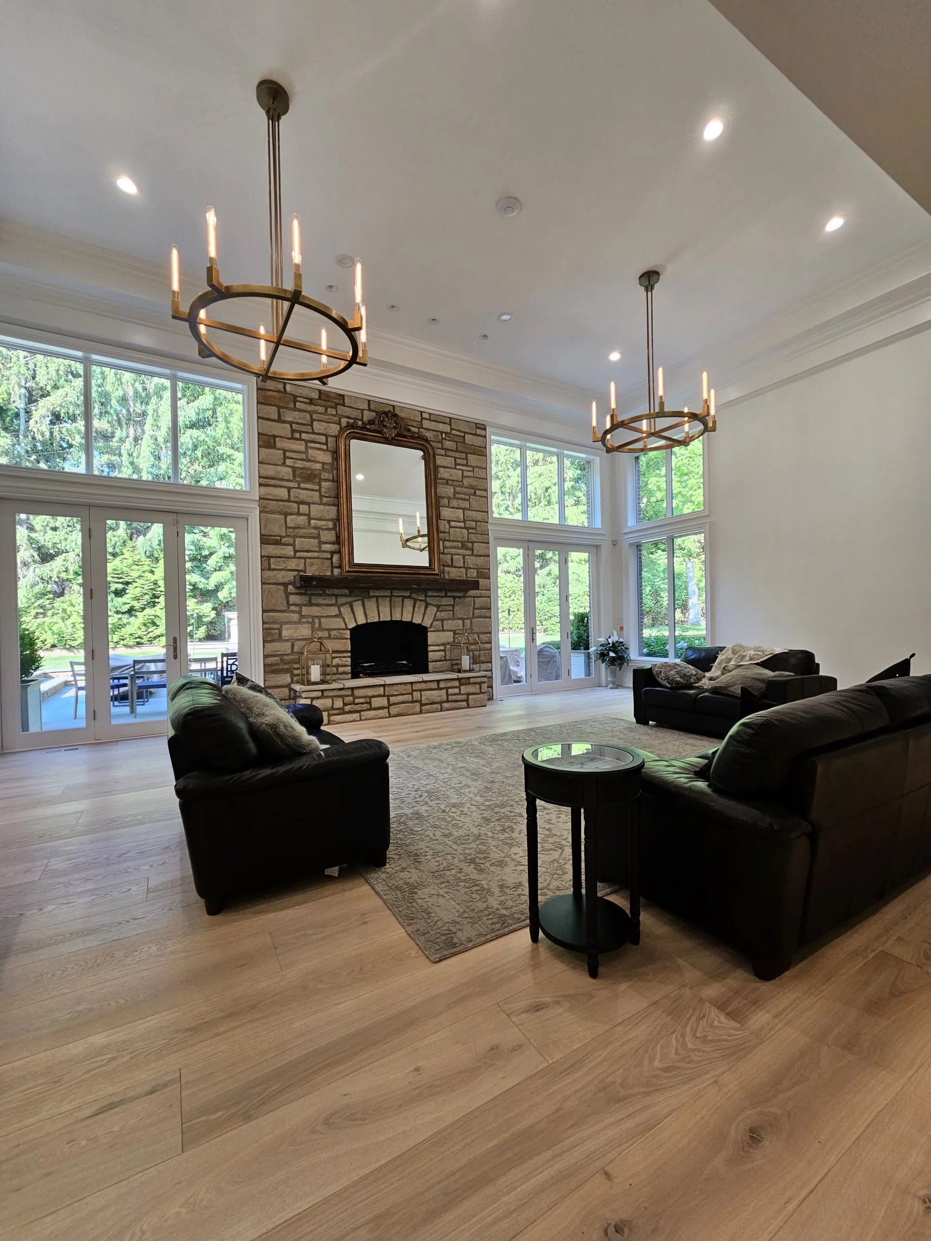 A living room with a brick fireplace, large windows, leather chairs, a matchstick chandelier, and a wooden floor.