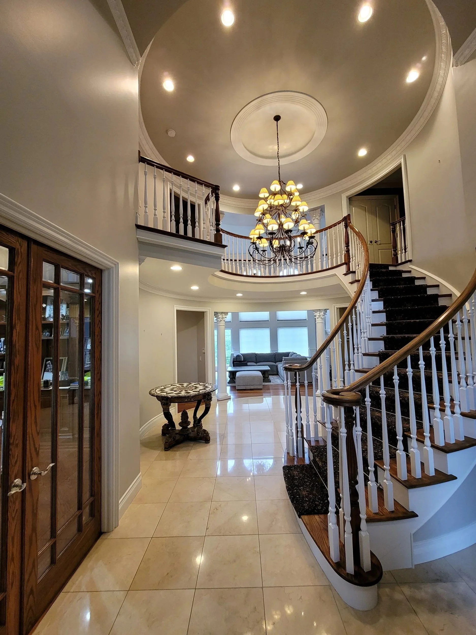 Interior view of a luxurious home with a grand staircase, chandelier, and sitting area near large windows, high ceiling with crown molding, tiled floor, and wooden accents.