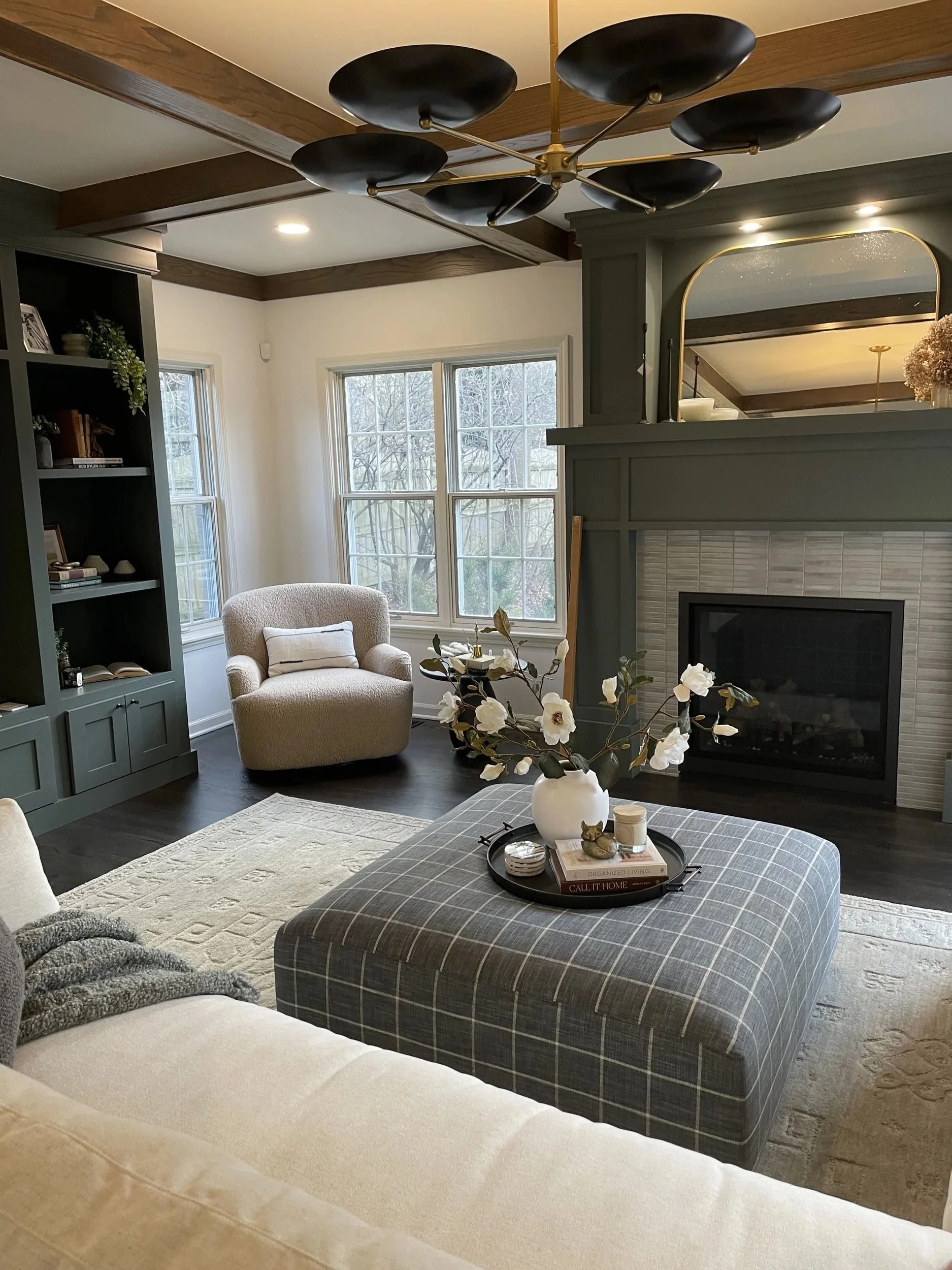 Living room with beige sofa, plaid ottoman, gray armchair, dark wood cabinet, fireplace, large window, modern chandelier.