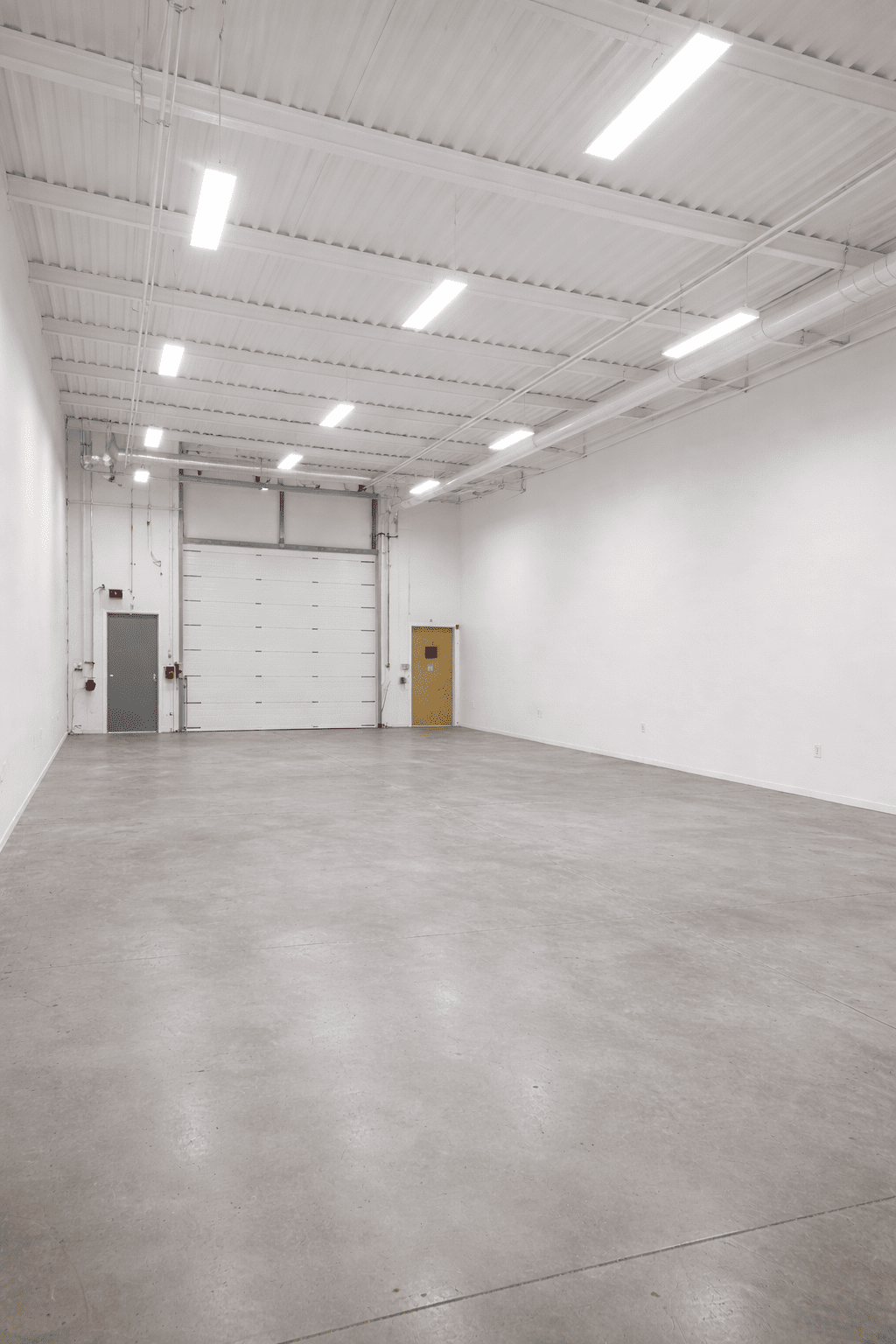 Empty industrial warehouse with a concrete floor, white walls, and ceiling with fluorescent lights. There is a large white garage door and two smaller doors, one gray and one yellow, along the back wall.