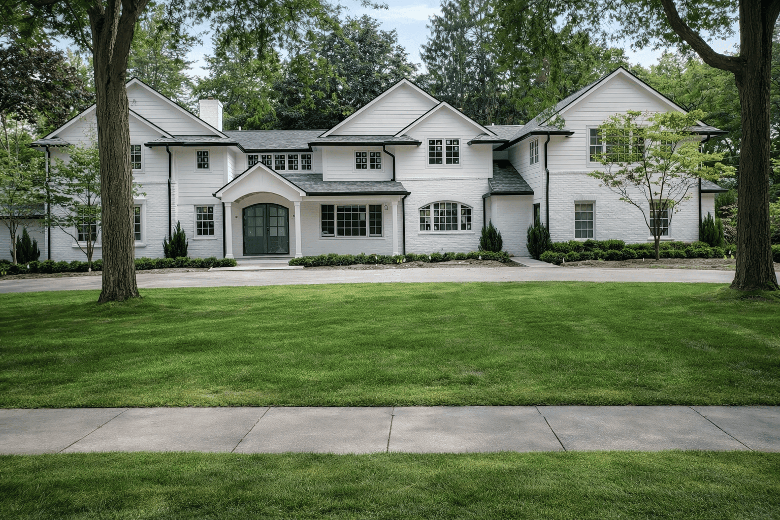 A large white house with multiple gabled roofs, surrounded by green trees and a well-maintained lawn, featuring a pathway leading to the front door.