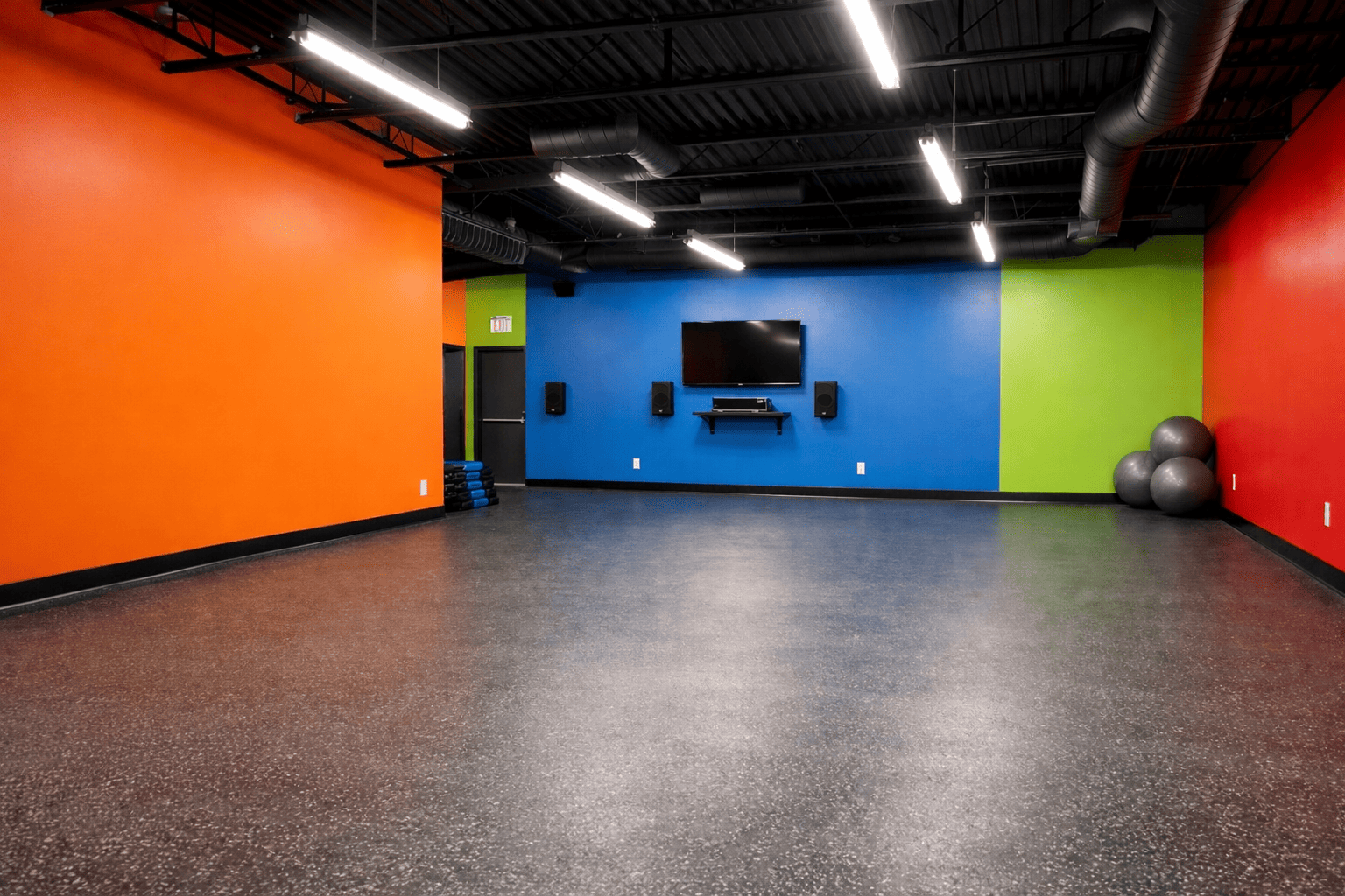 Empty gym or fitness room with colorful walls, a wall-mounted TV, exercise balls, and workout mats.