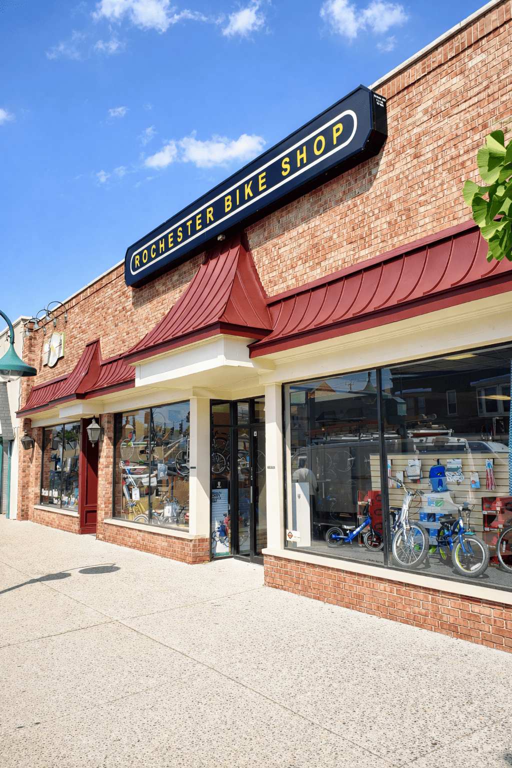 Exterior of Rochester Bike Shop with large windows displaying bicycles and biking accessories, brick facade, red metal awnings, and a sign overhead reading 'Rochester Bike Shop'.