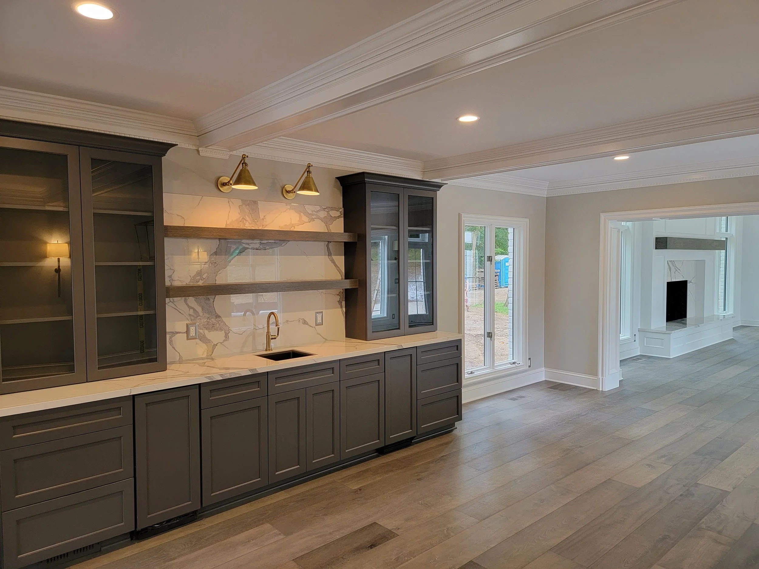Modern kitchen with gray cabinetry, marble backsplash, open shelving, and built-in glass cabinets, leading to a living room with fireplace and large windows.