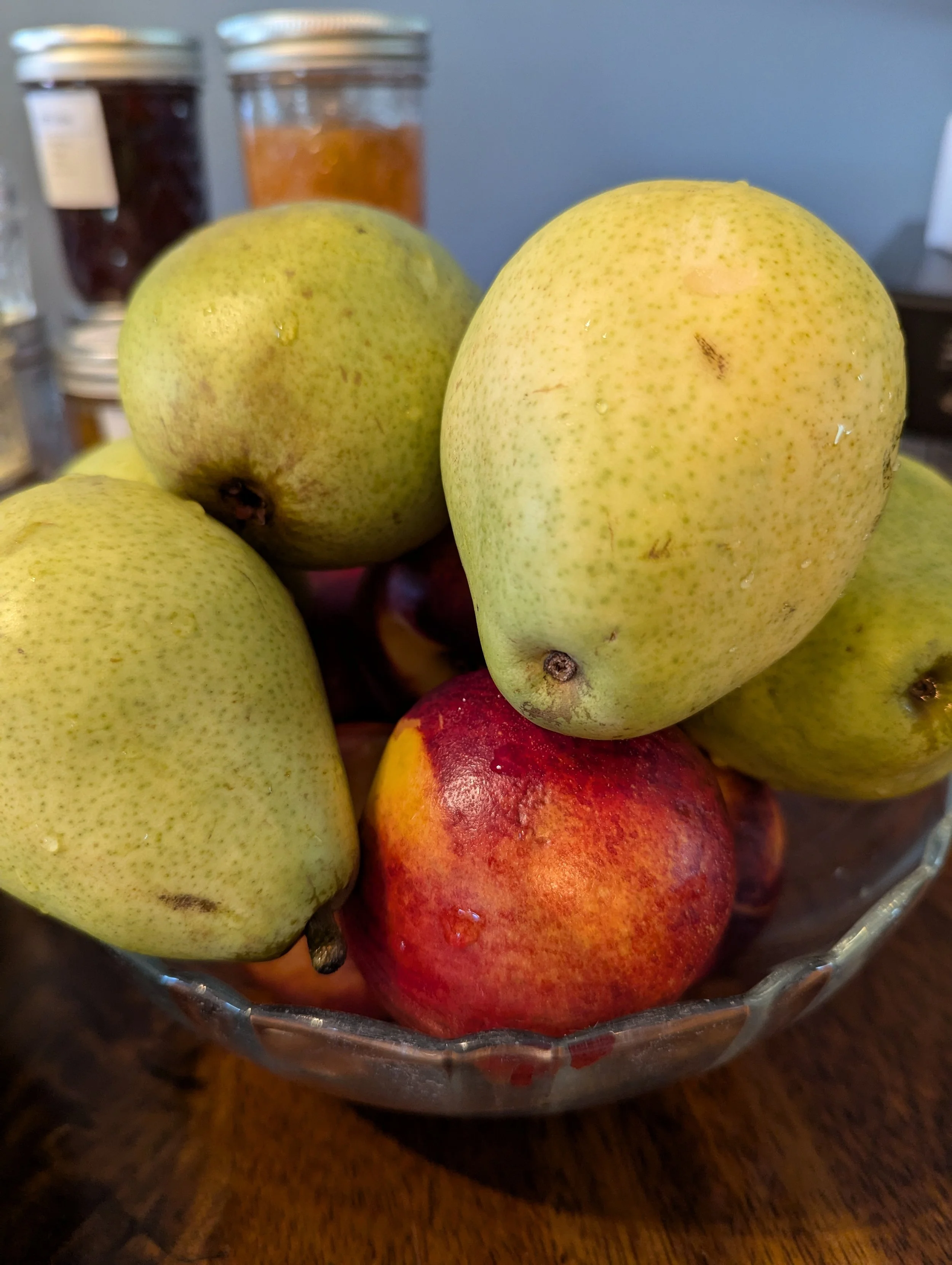 A glass bowl filled with pears and apples, with pears being yellowish-green with small brown spots and apples being red with some yellow patches.