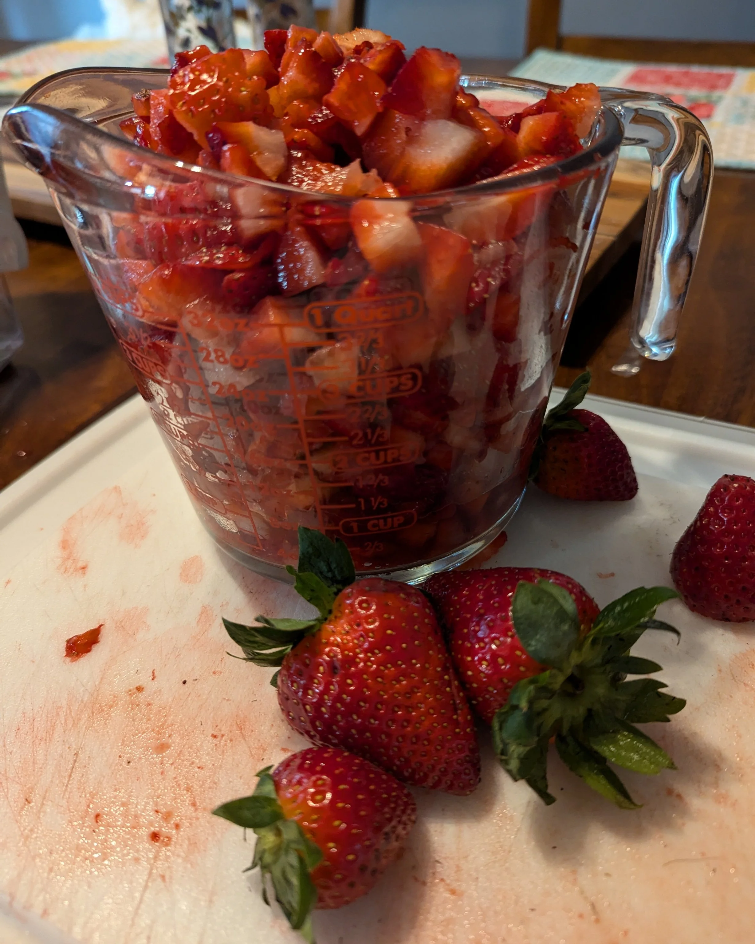 A glass measuring cup filled with chopped strawberries on a white cutting board with whole strawberries nearby.