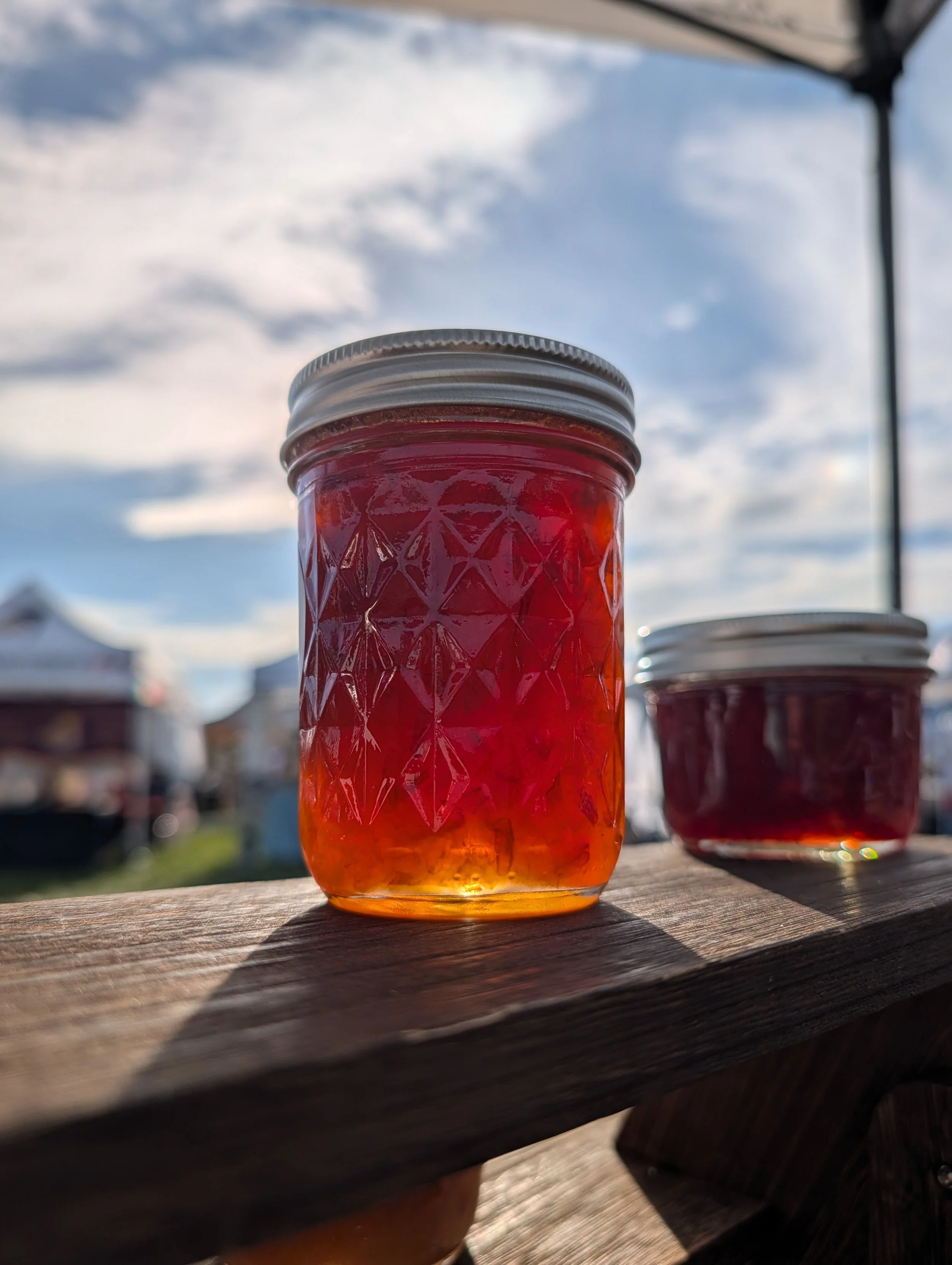 A close-up of a mason jar filled with red jelly on a wooden surface outdoors, with a cloudy sky in the background.