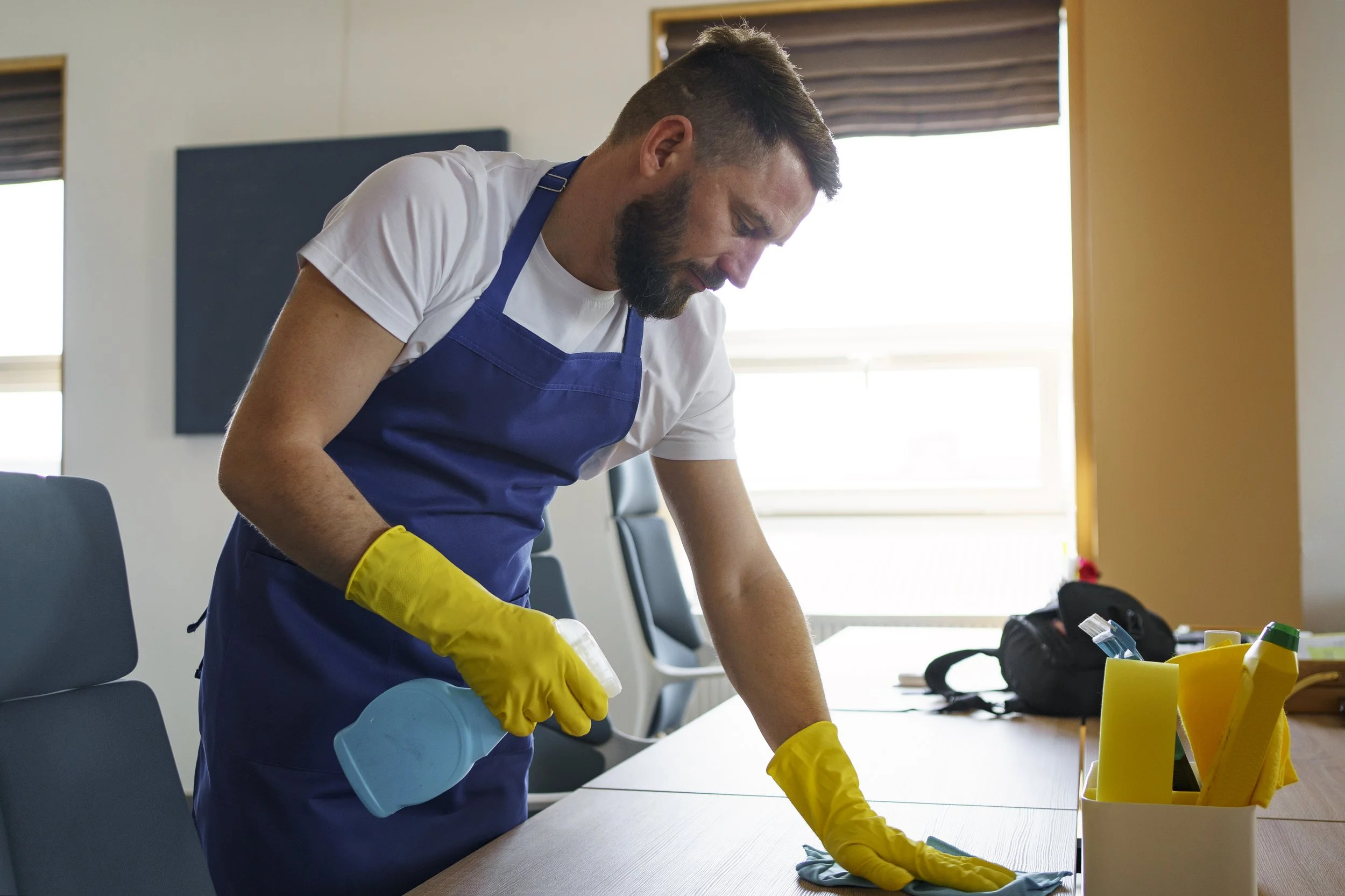 Man in a white t-shirt and blue apron cleaning a conference table with yellow gloves, using a spray bottle and cloth, in a room with sunlight coming through the windows.
