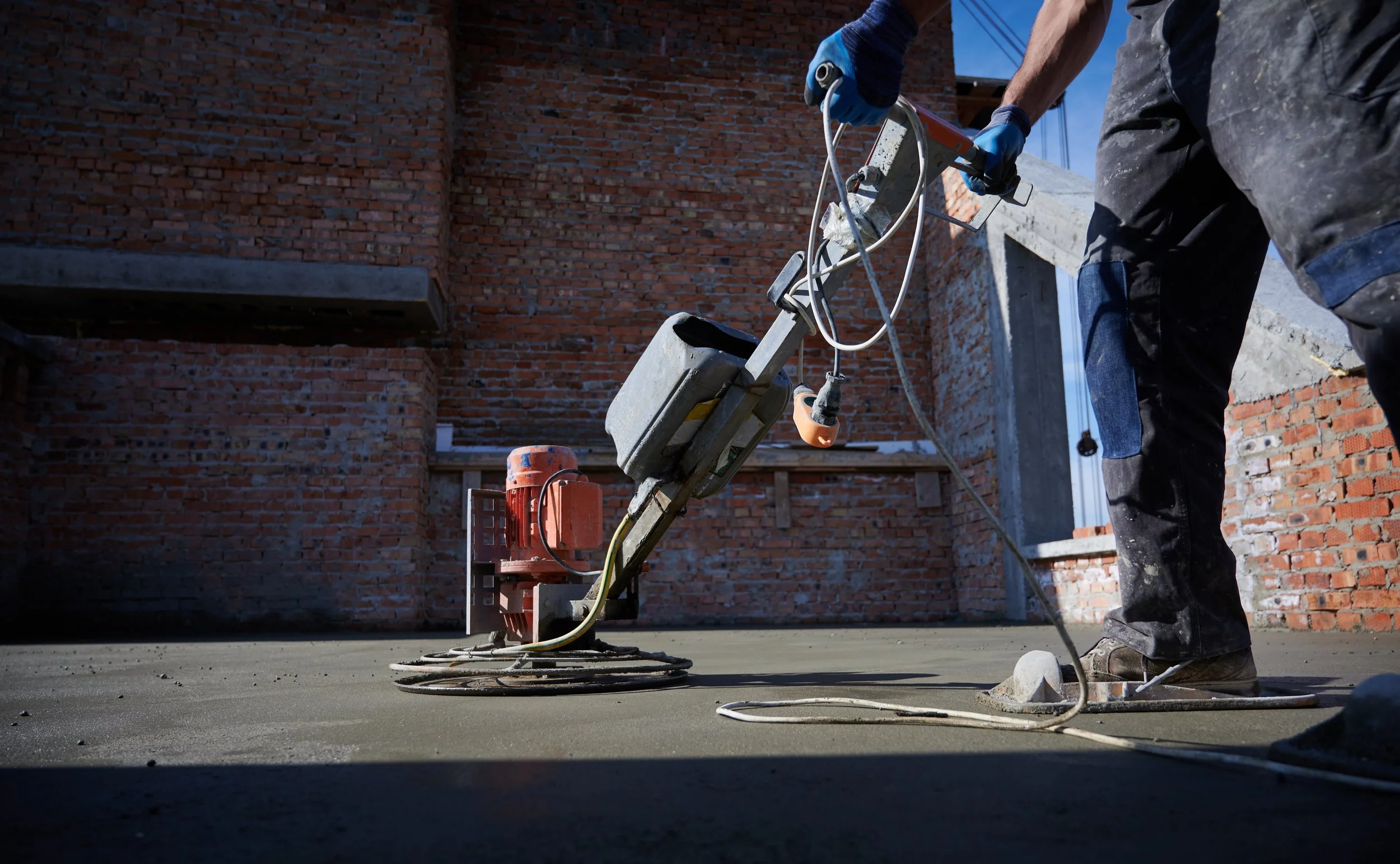 Construction worker using a power screed to level concrete on a building site with brick walls in the background.