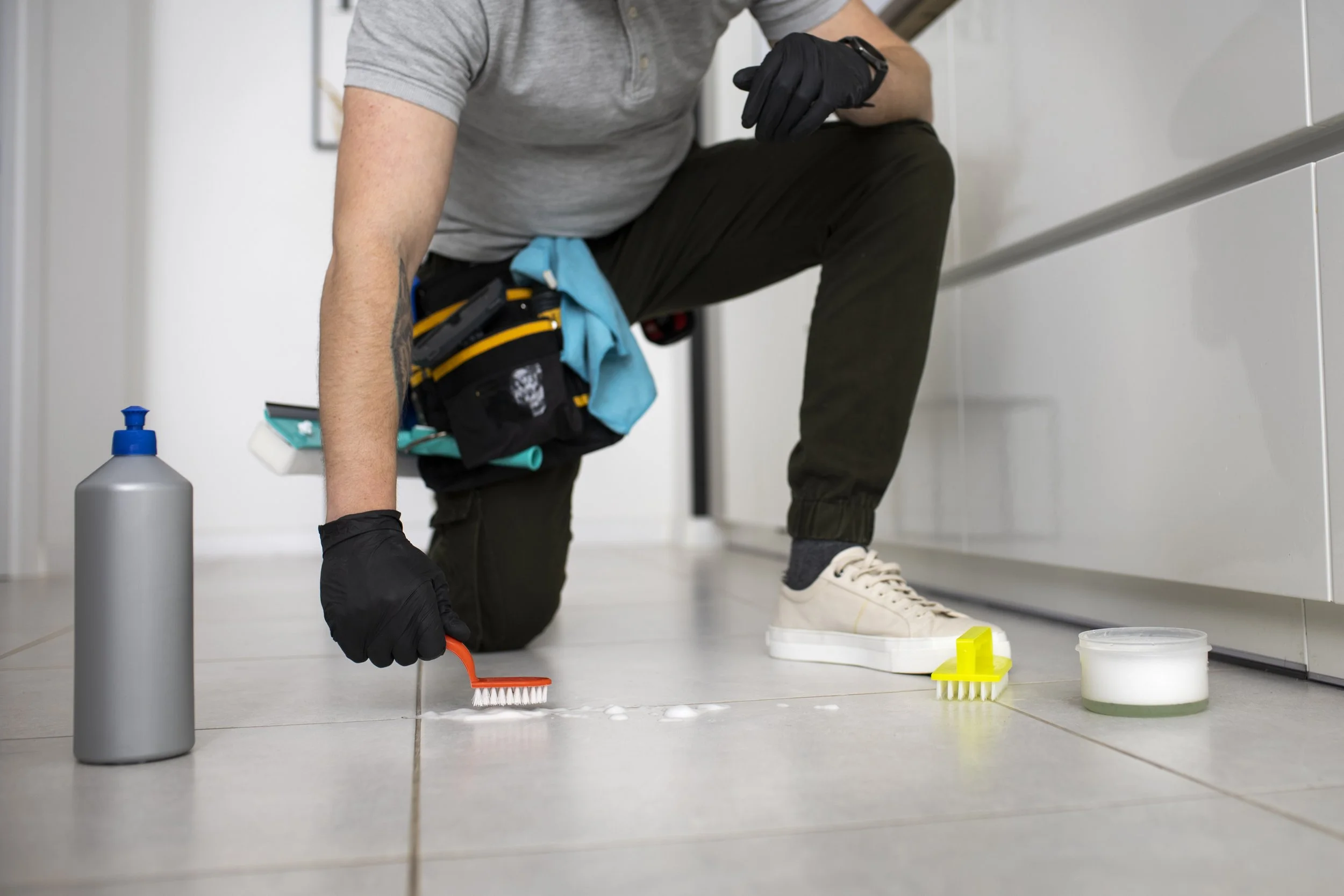 Person in casual clothing and black gloves cleaning a tiled floor with brushes and cleaning supplies, including a bottle and a container of cleaning solution.