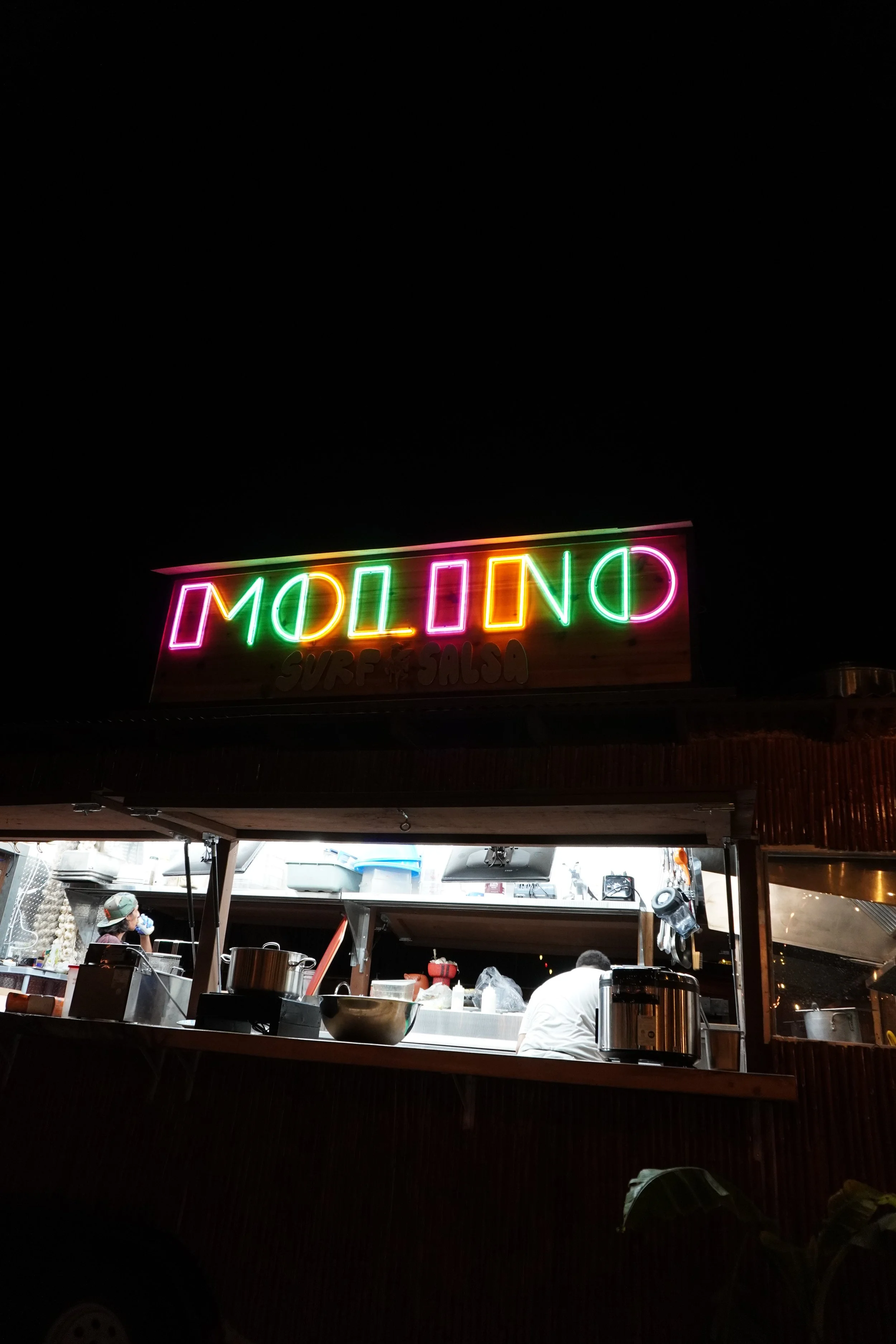 Nighttime view of a food stand with a neon sign that spells 'MOLINO' in colorful lights, and a smaller sign below reading 'SURF SALOON'. The stand has two people working inside, with various kitchen equipment and supplies visible.