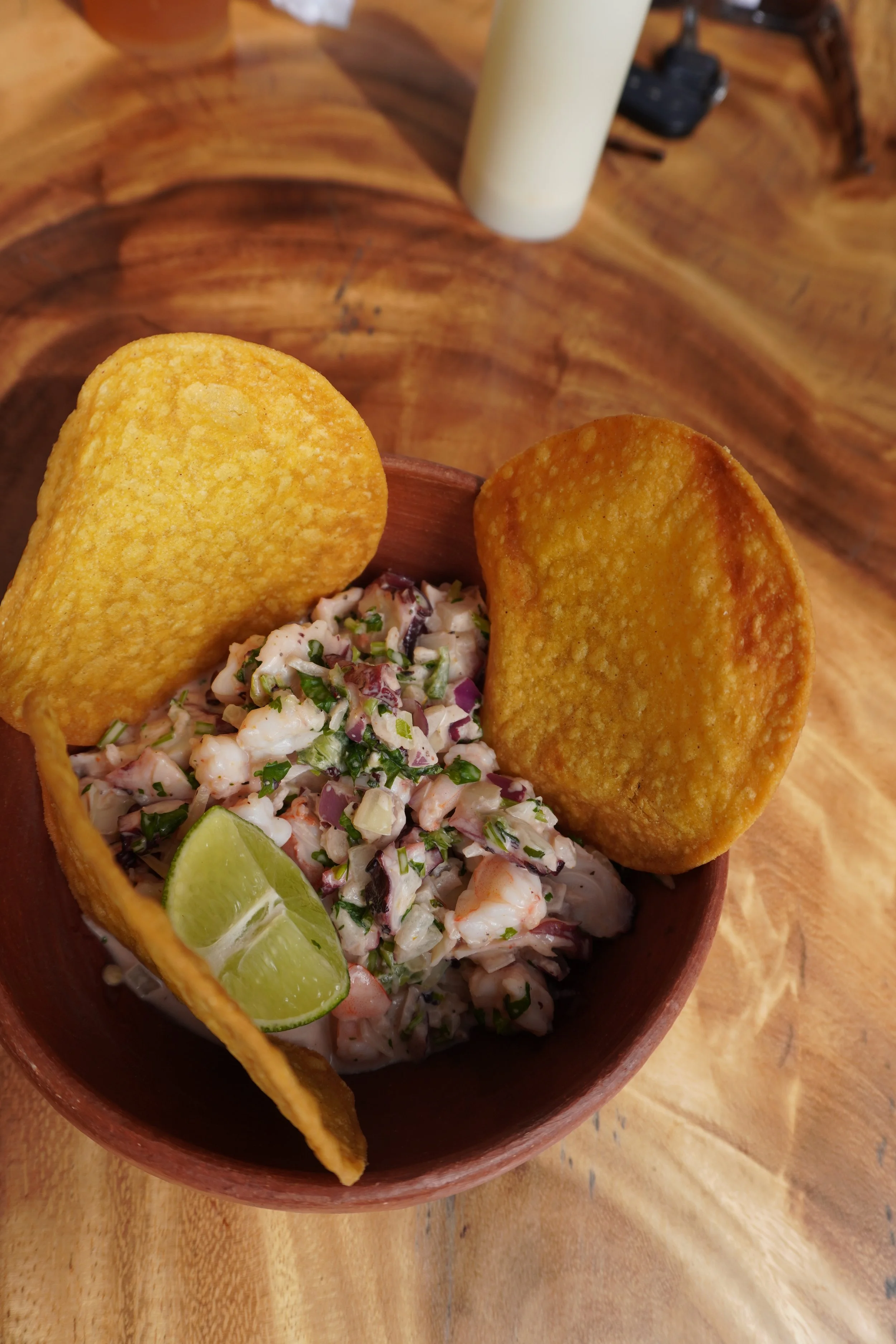 A wooden bowl containing shrimp ceviche with chopped vegetables, garnished with lime wedges, and two large tortilla chips on top, on a wooden table with a candle and other objects in the background.