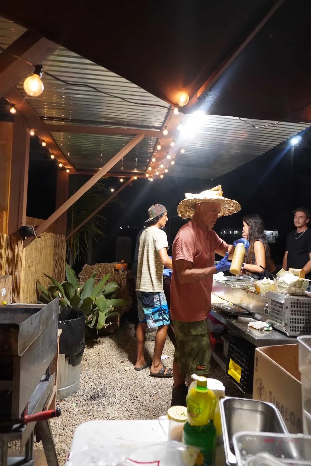 People preparing food at an outdoor night market or food stall, with string lights overhead, a man wearing a large straw hat, and others in casual attire.
