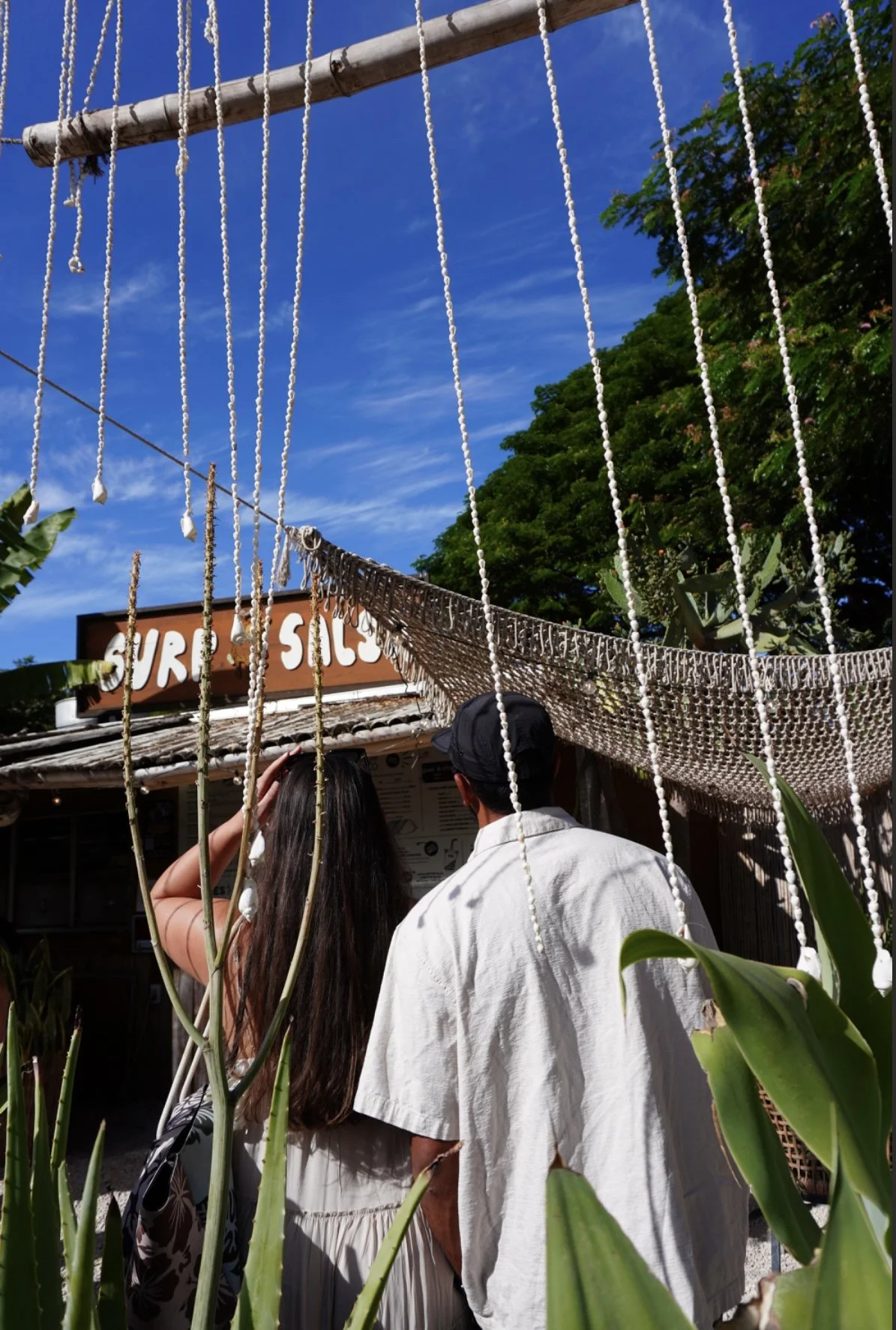 Two people standing in line outside a burger and taco food stand with a sign that says 'BURGER' and 'TACOS' on a sunny day with blue skies and lush green trees.