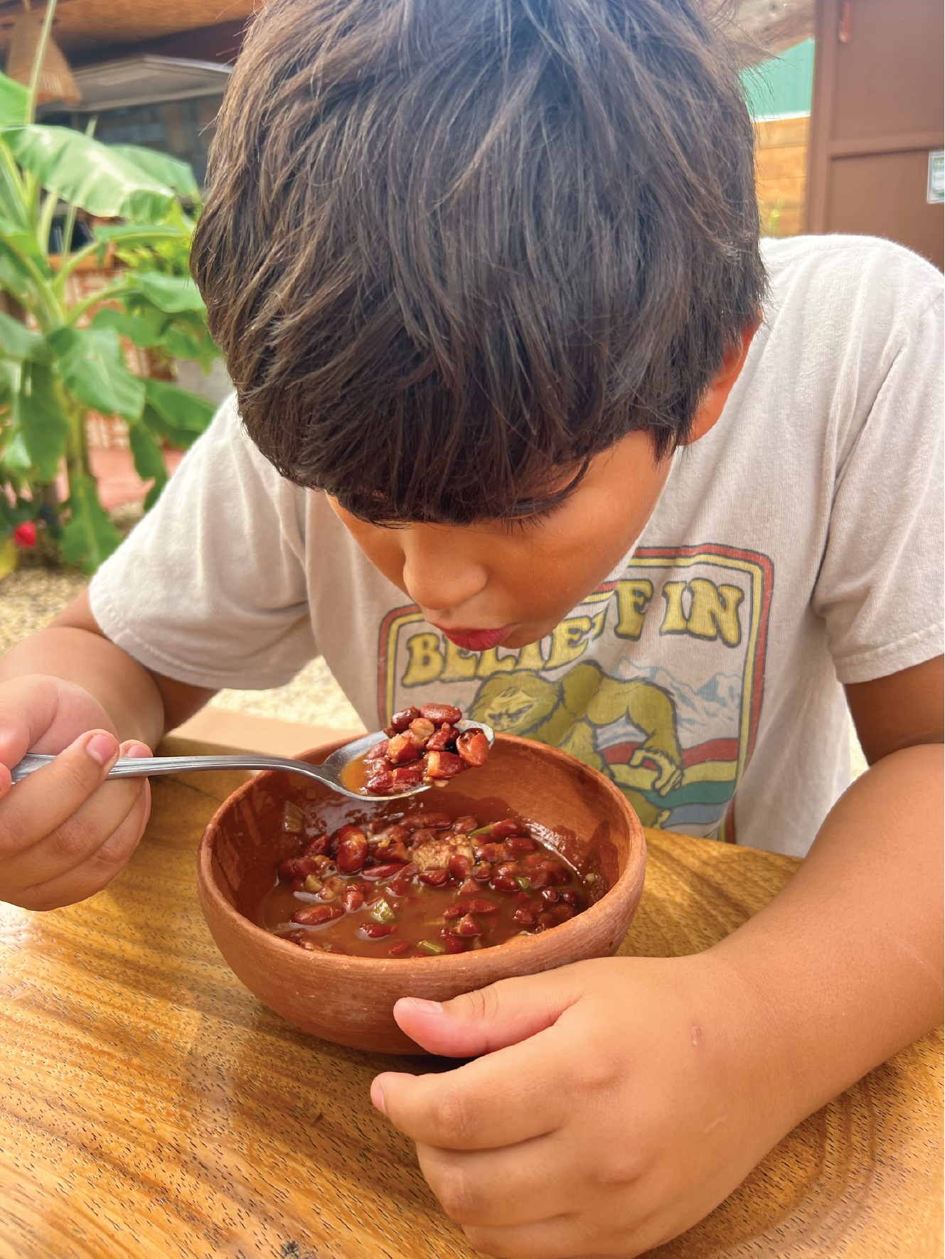 A young boy with dark brown hair eating a bowl of chili with beans, using a spoon, sitting at a wooden table outdoors.