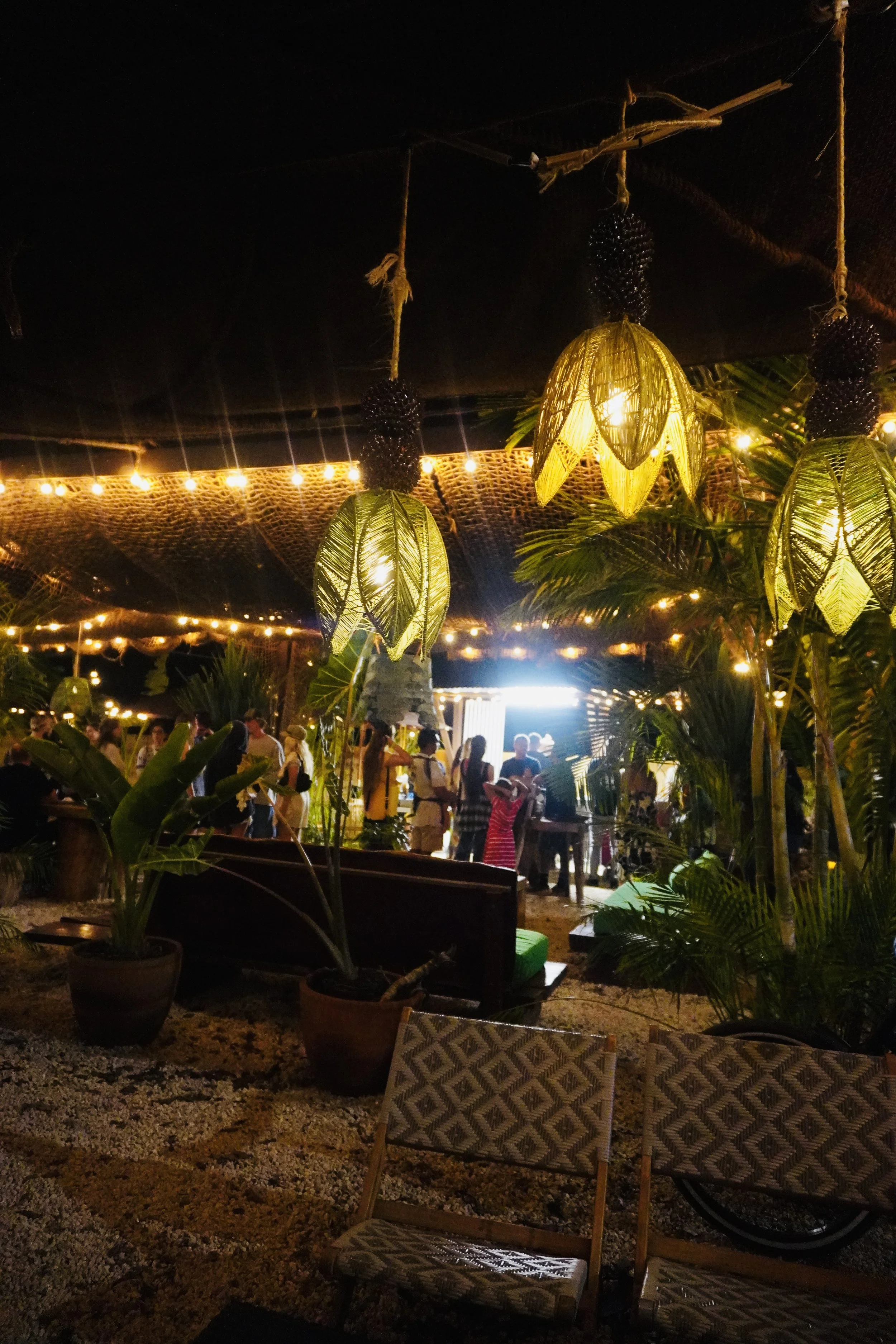 Nighttime outdoor gathering with hanging lantern lights, tropical plants, and a group of people socializing at a decorated venue or patio.