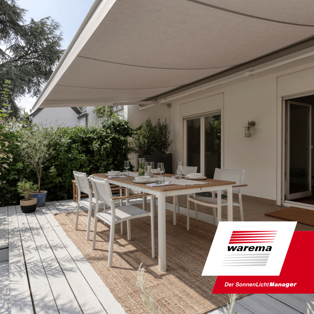 A sunlit outdoor patio with a white dining table set with white plates, bowls, and glassware, surrounded by white chairs, adjacent to a house with sliding glass doors, shaded by a large retractable beige awning, with green plants and trees in the bac