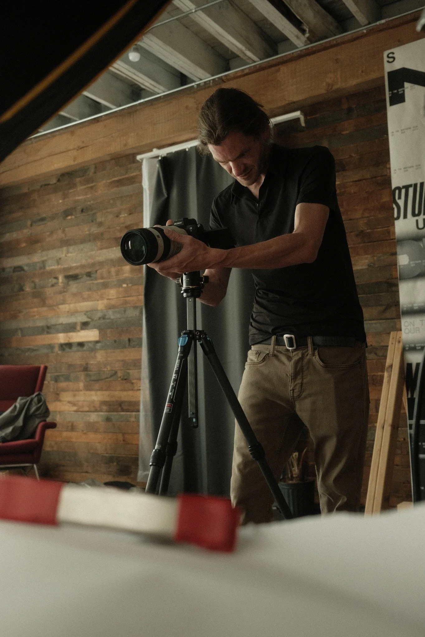 A man with long hair, wearing a black shirt and brown pants, is adjusting a camera on a tripod in a room with wood-paneled walls.