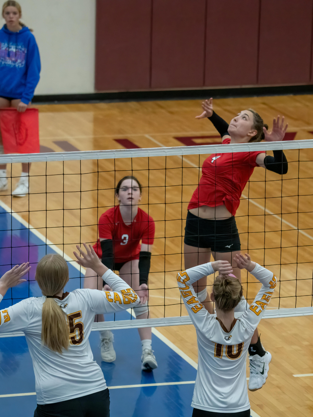 A volleyball game in progress with two players in white jerseys at the net blocking, and a player in a red jersey jumping to hit the ball over the net. Another player in a red jersey is crouching behind the hitter, observing the play, on a wooden gym floor.