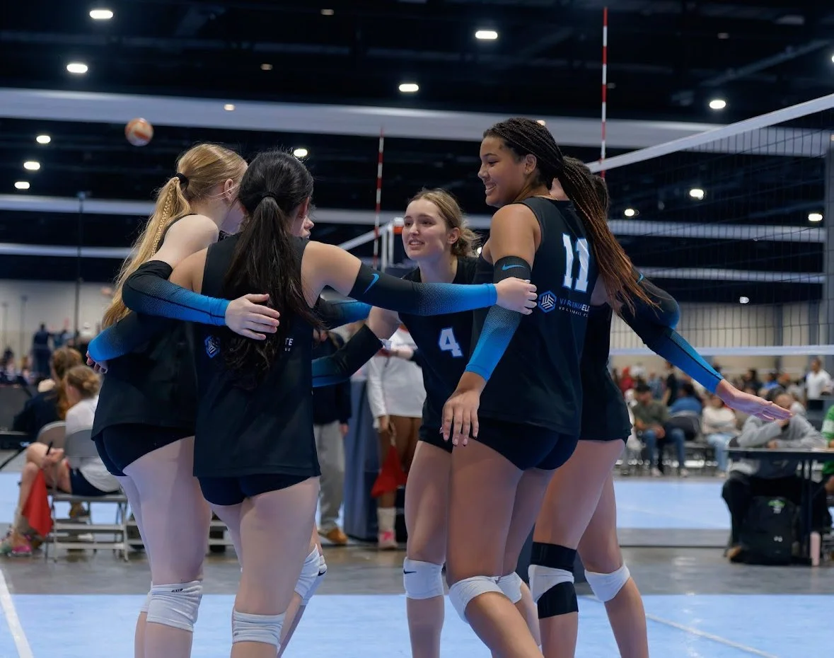 A group of five female volleyball players in black uniforms celebrating on an indoor court during a match.
