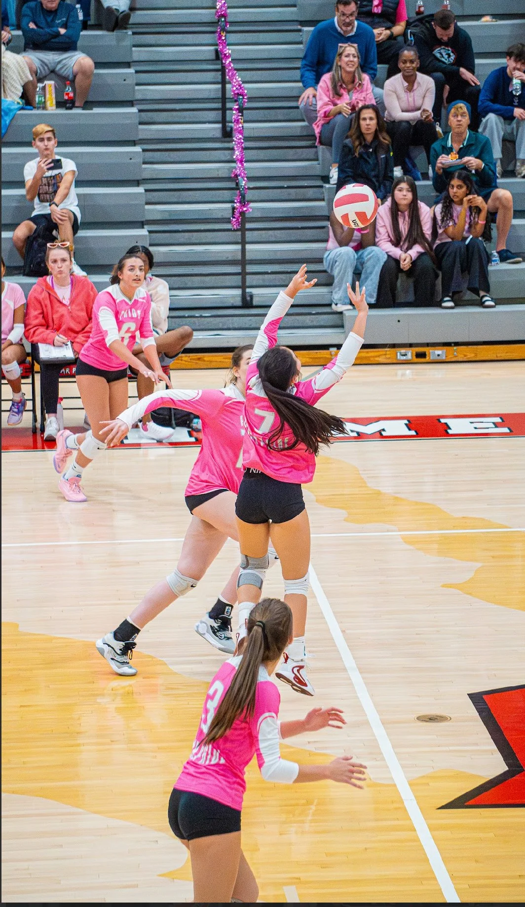 Girls volleyball team in pink uniforms playing on the court during a match, with an audience seated in the bleachers.