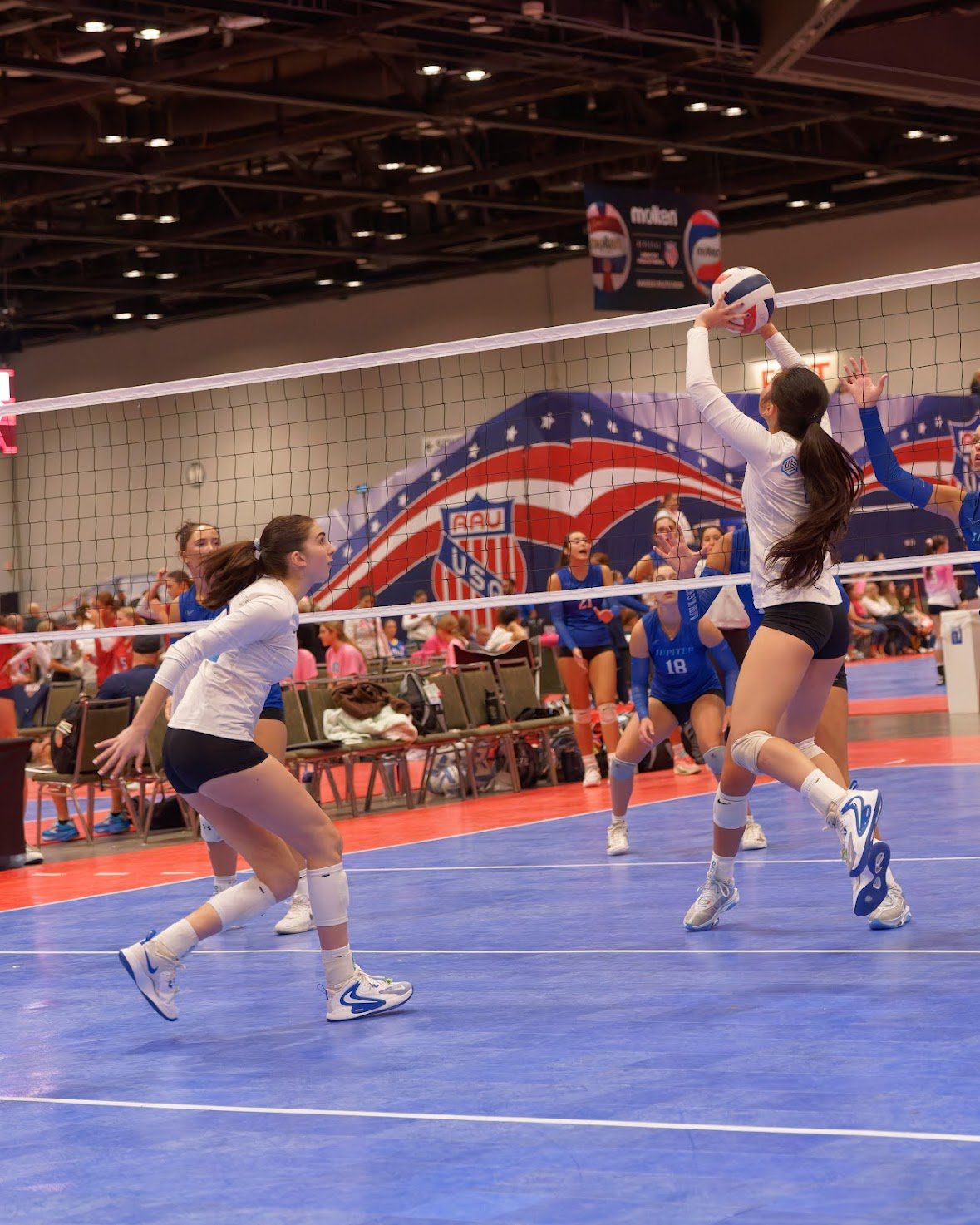 Girls playing volleyball in an indoor gym, with a volleyball net and an American flag-themed banner in the background.