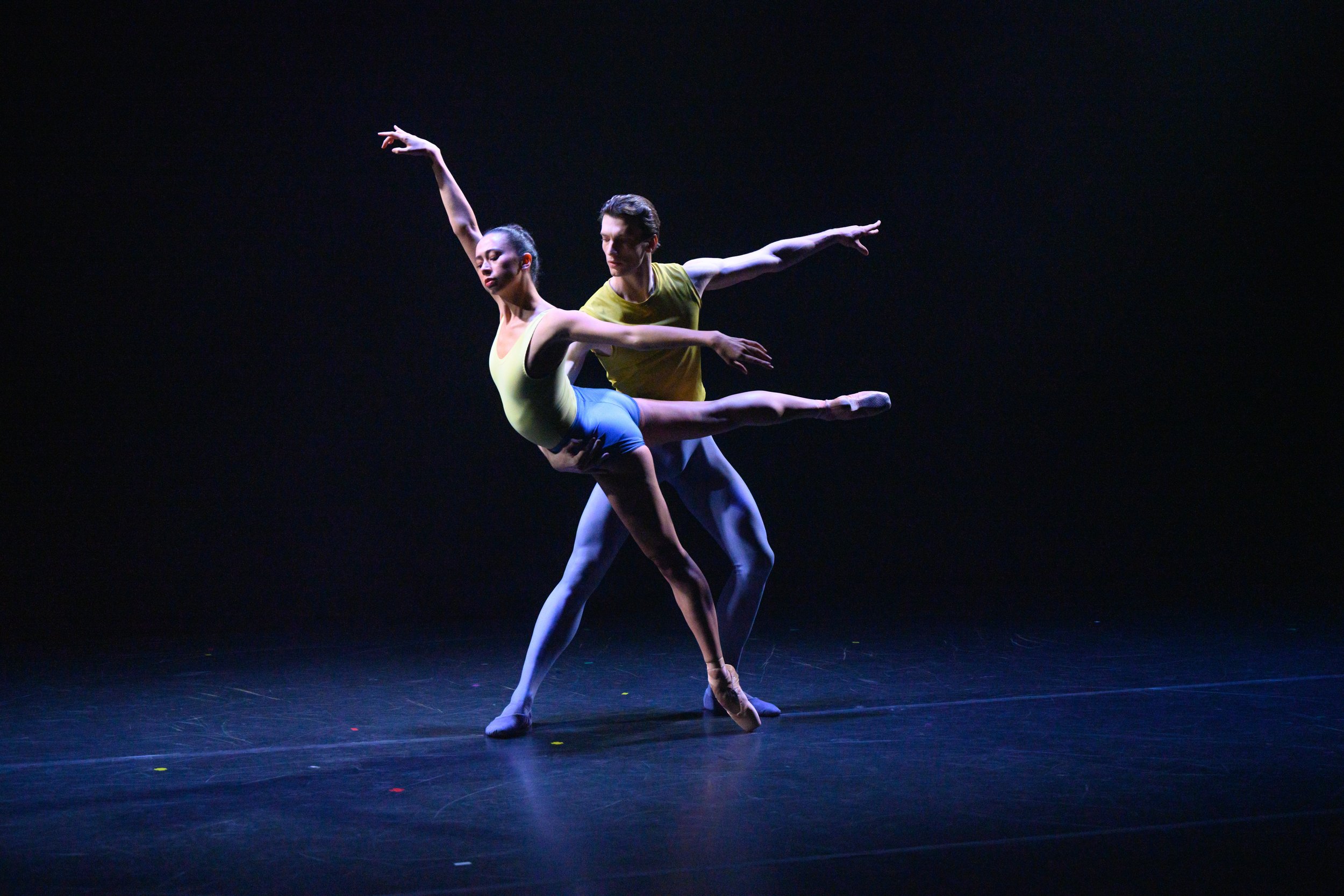 Two ballet dancers perform on stage with a dark background, one dancer in a yellow top and blue bottoms, the other in a light yellow leotard, both in extended dance poses.