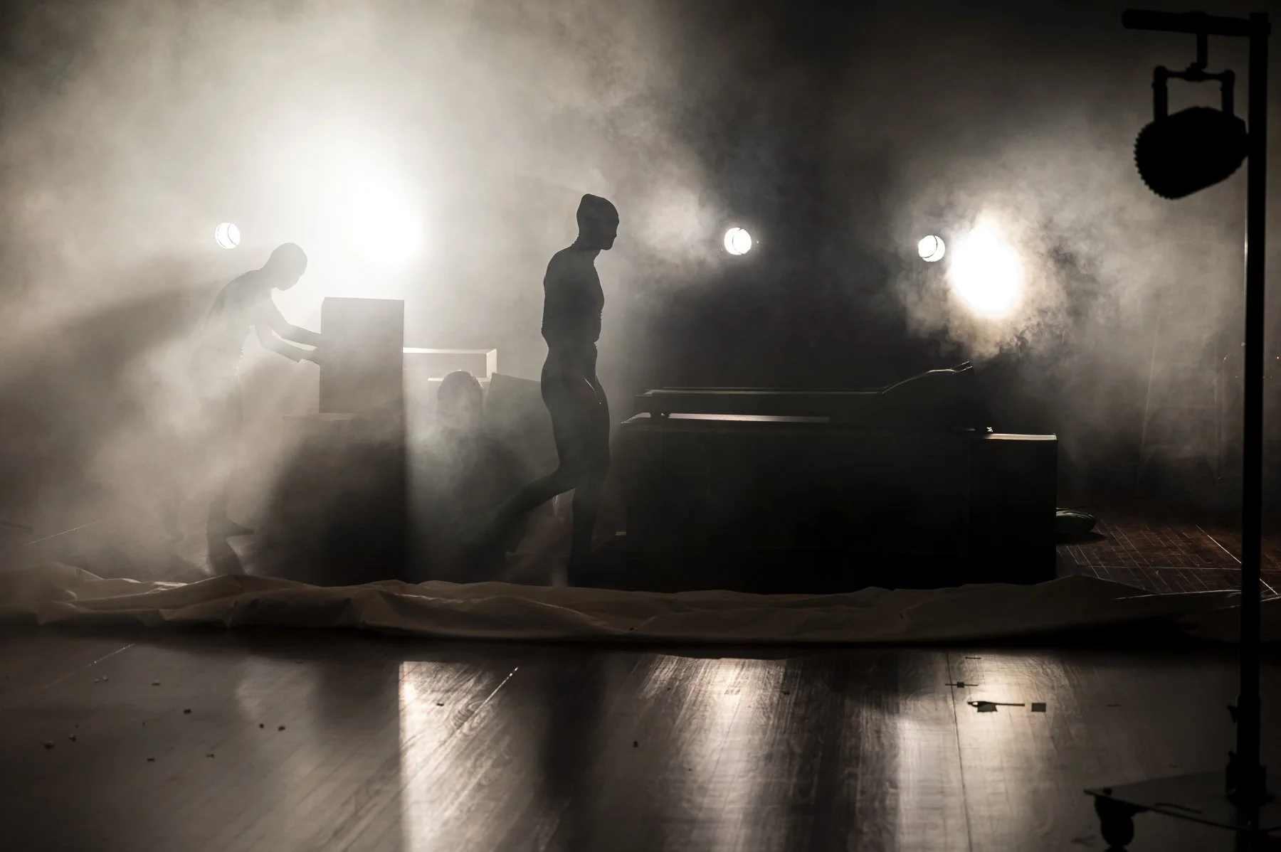 Silhouettes of two people working on a stage or set, surrounded by mist and backlit with bright lights, with a wooden floor and equipment visible.