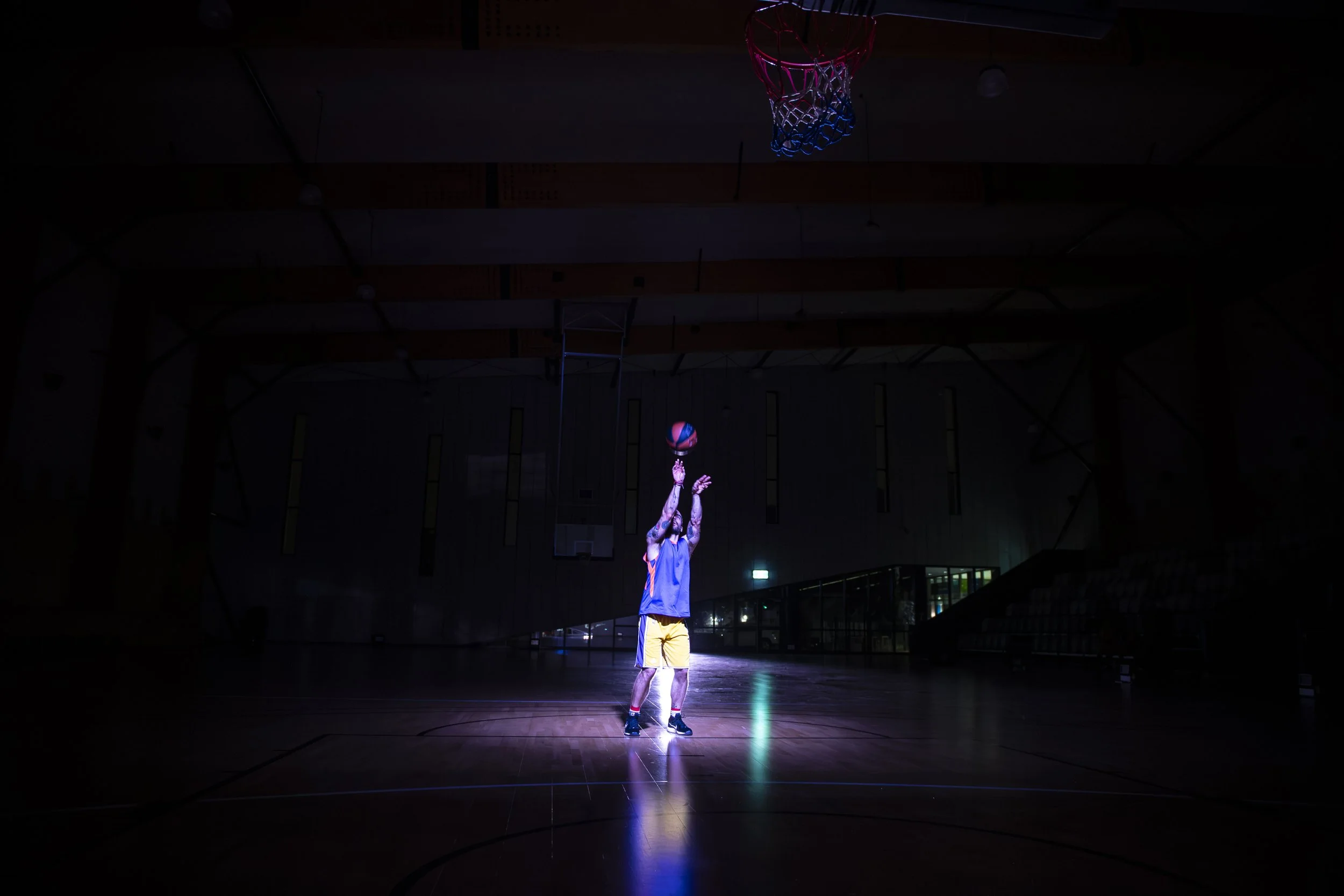 A basketball player in a yellow and blue uniform taking a shot in a dimly lit basketball court with a bright spotlight on him.