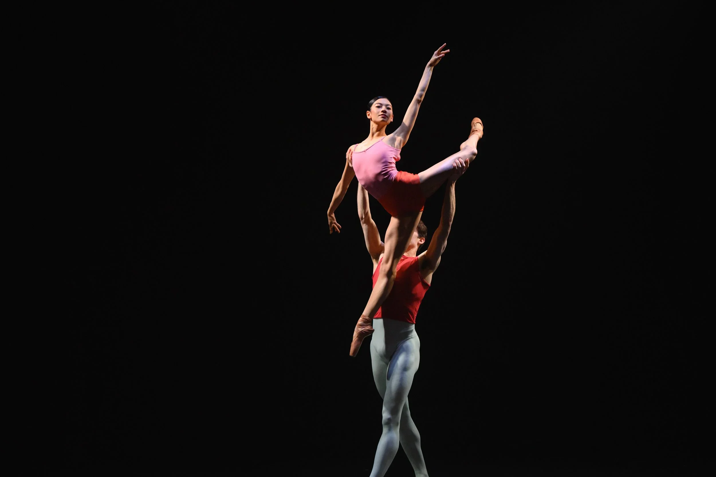 Two ballet dancers perform an acrobatic pose on stage with a dark background.