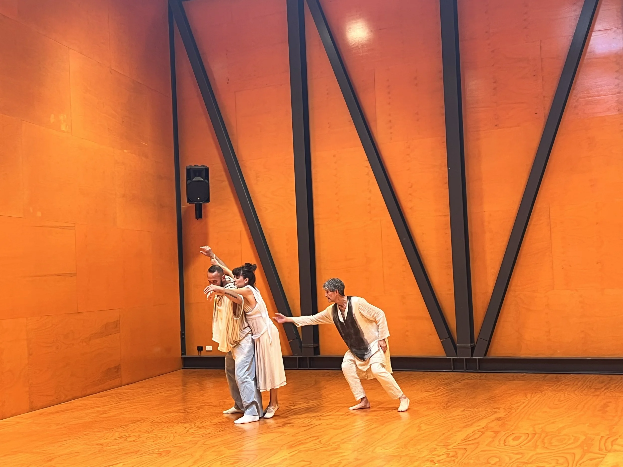 Three dancers perform in a room with orange wooden walls and floor, with black structural beams and a speaker on the wall.