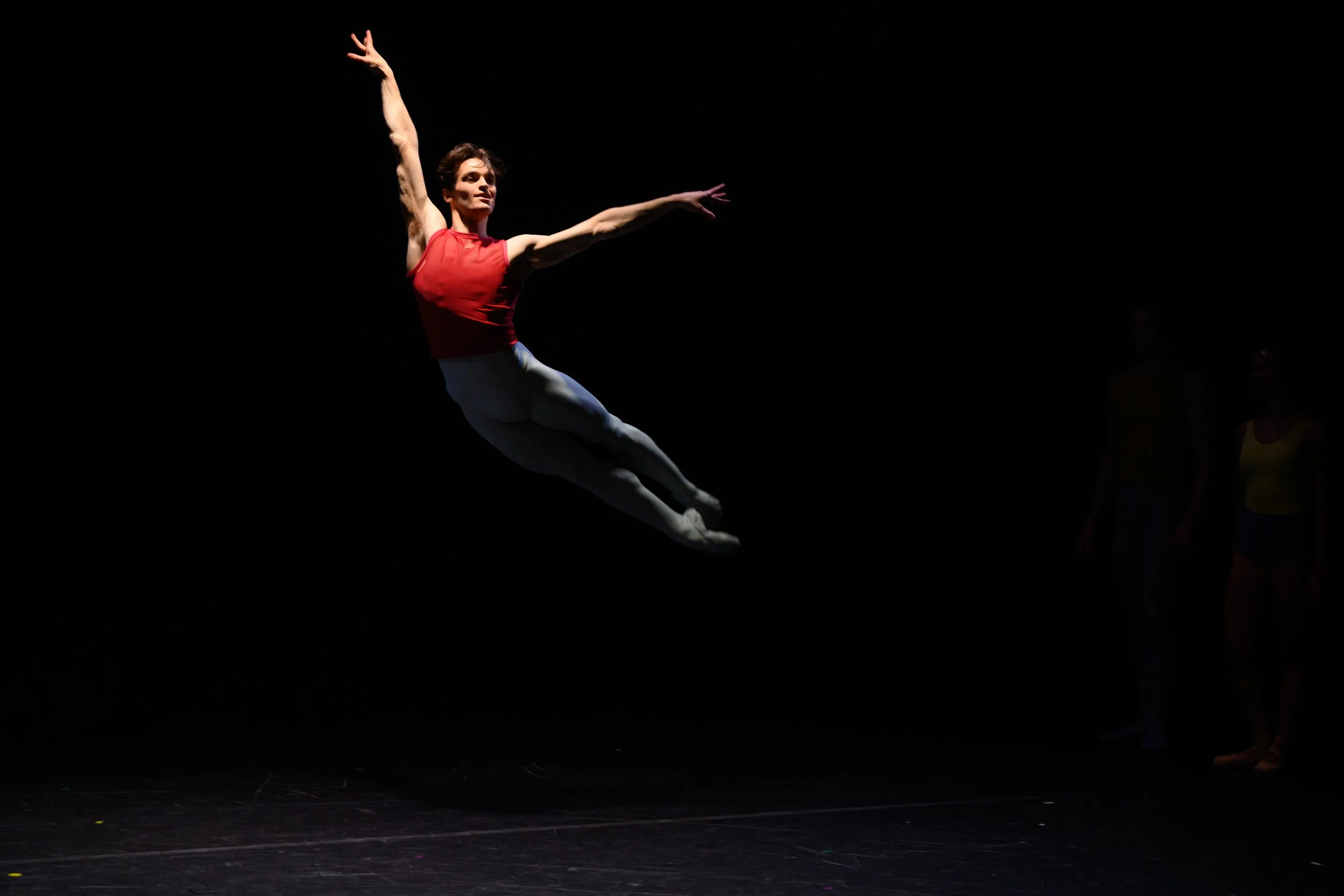 A female performer in mid-air on a dark stage, wearing a red top and gray leggings, with two people in the background.