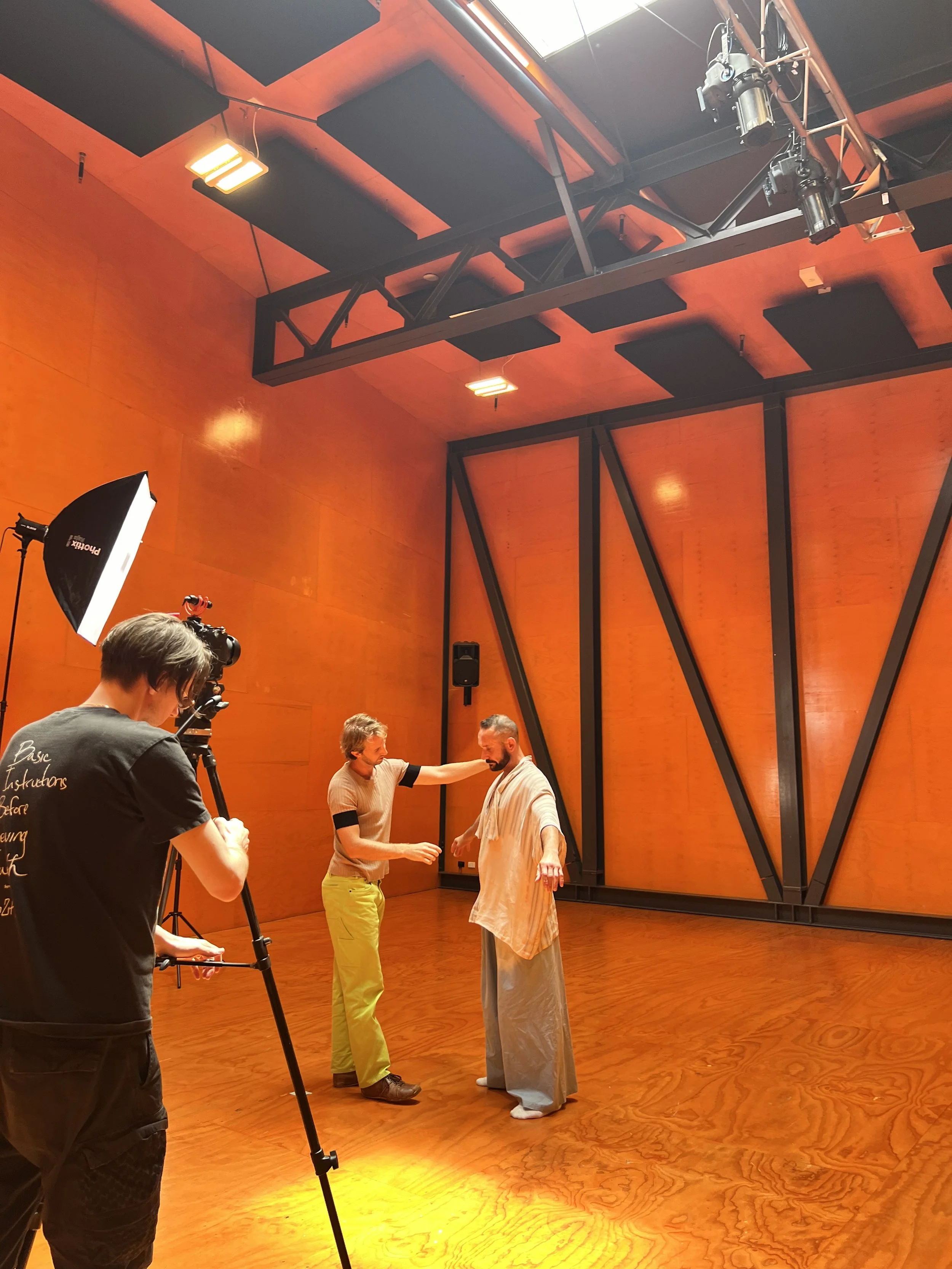 A film crew is recording a scene in a wooden studio with orange walls. Two men are in the center, one touching the other's chin while the other has a shawl, and a cameraman is filming them.