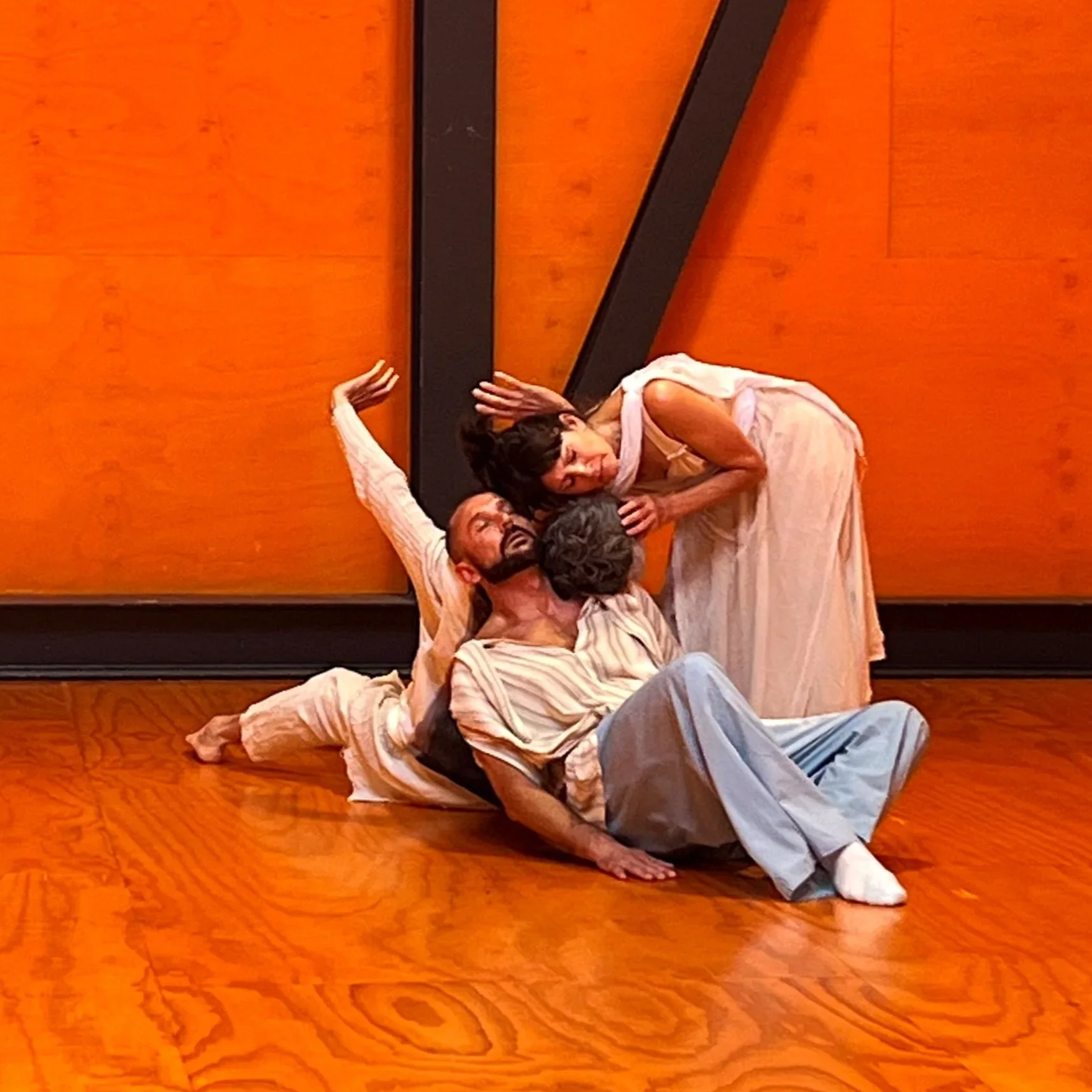 Four performers in period costumes perform a theatrical scene on a wooden stage with orange walls, with one lying on the floor, one sitting, and two leaning over each other in expressive poses.
