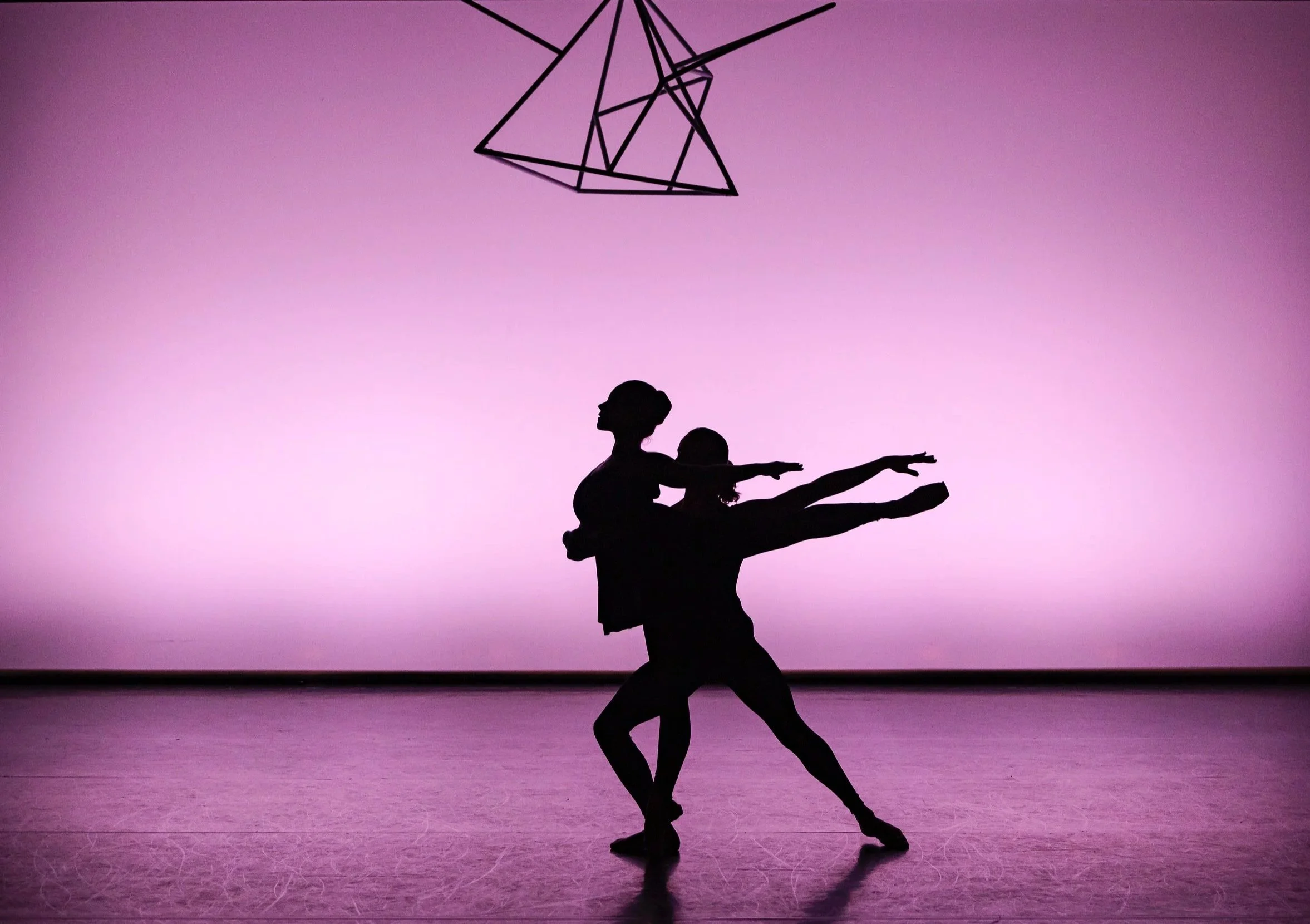 Silhouette of two ballet dancers, one adult and one child, performing on stage with a purple background and a geometric light fixture overhead.