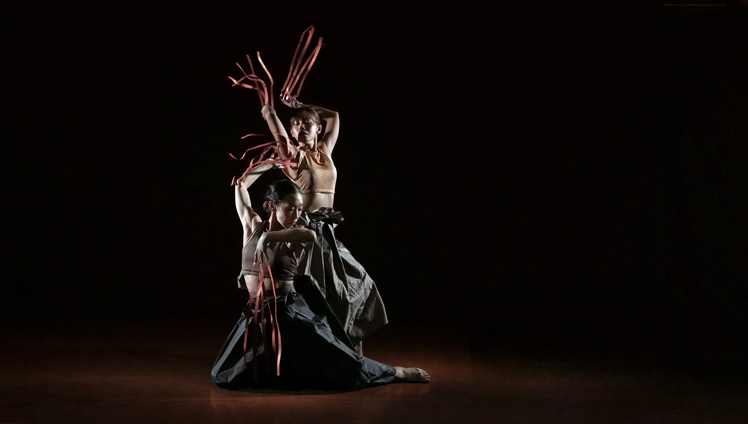 Two female dancers in dark clothing perform an intricate dance on a dark stage with minimal lighting, one standing with arms raised and the other kneeling with her eyes closed.